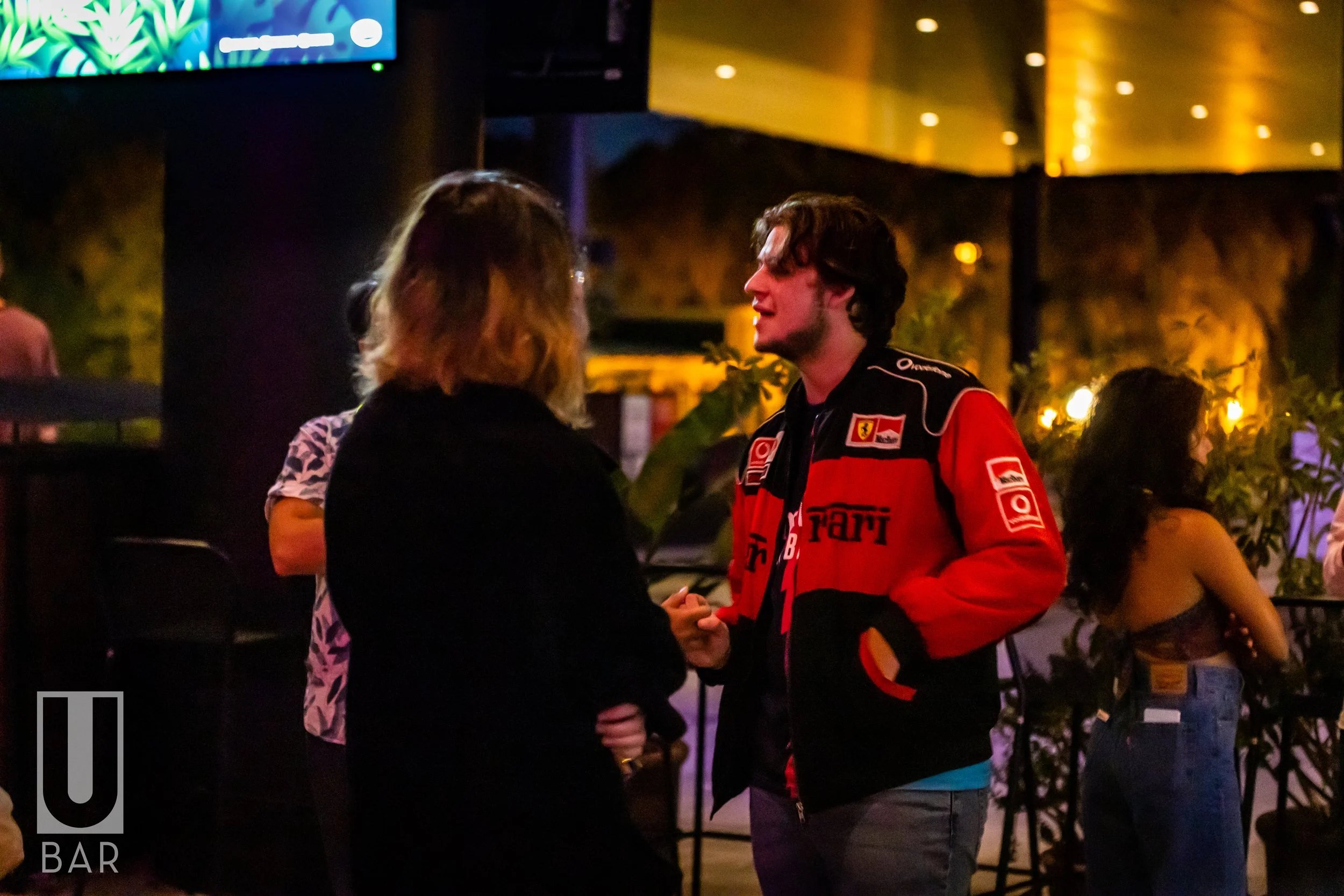 Two people engaged in conversation in a dimly lit bar or lounge with warm lighting. One is wearing a Ferrari racing jacket, and the other is dressed in darker clothing. There are other patrons in the background, with some sitting and others standing,