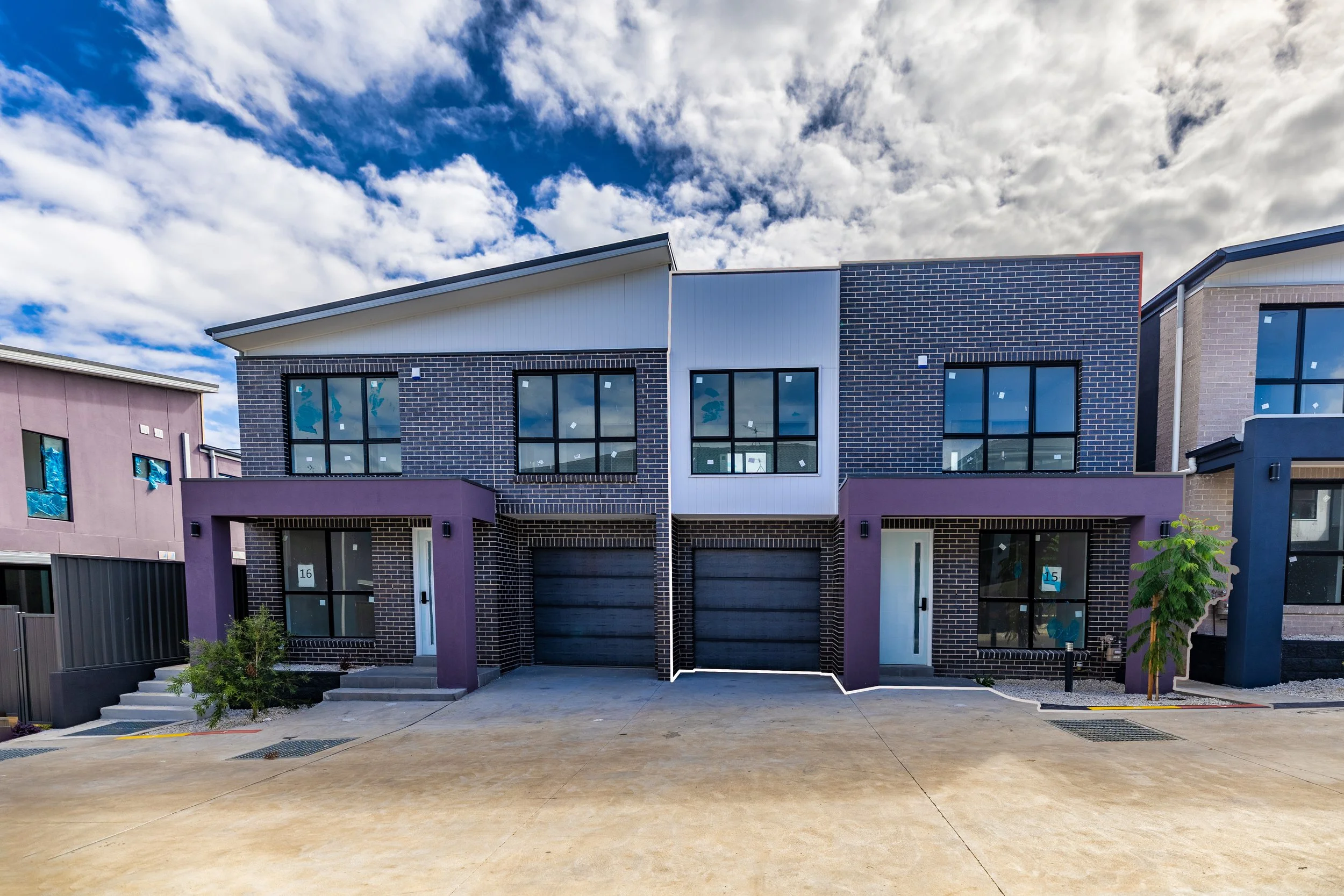 Front view of new modern townhouse with black brick exterior, large upper windows, and garage doors, under partly cloudy sky.