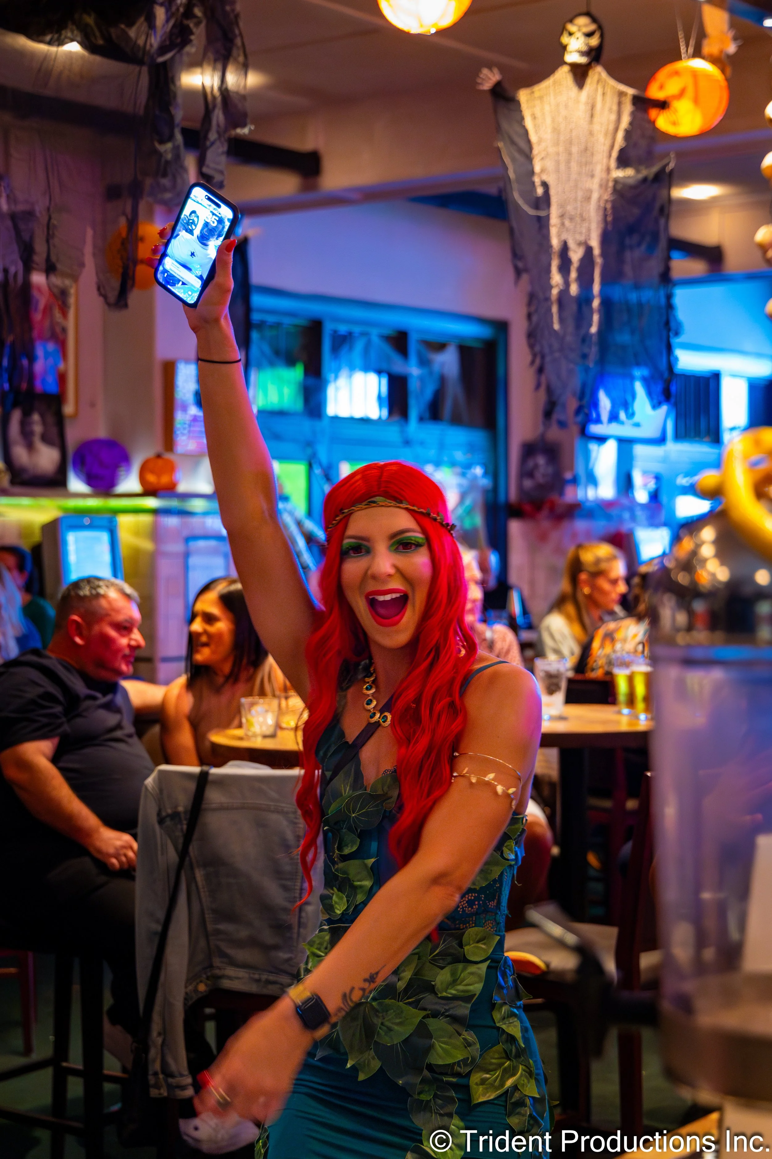 A woman with long red hair, wearing a leaf-patterned dress and jewelry, is standing in a decorated room holding up a smartphone, smiling excitedly for a selfie. The room has Halloween decorations including hanging ghosts, pumpkins, and skeleton figures.