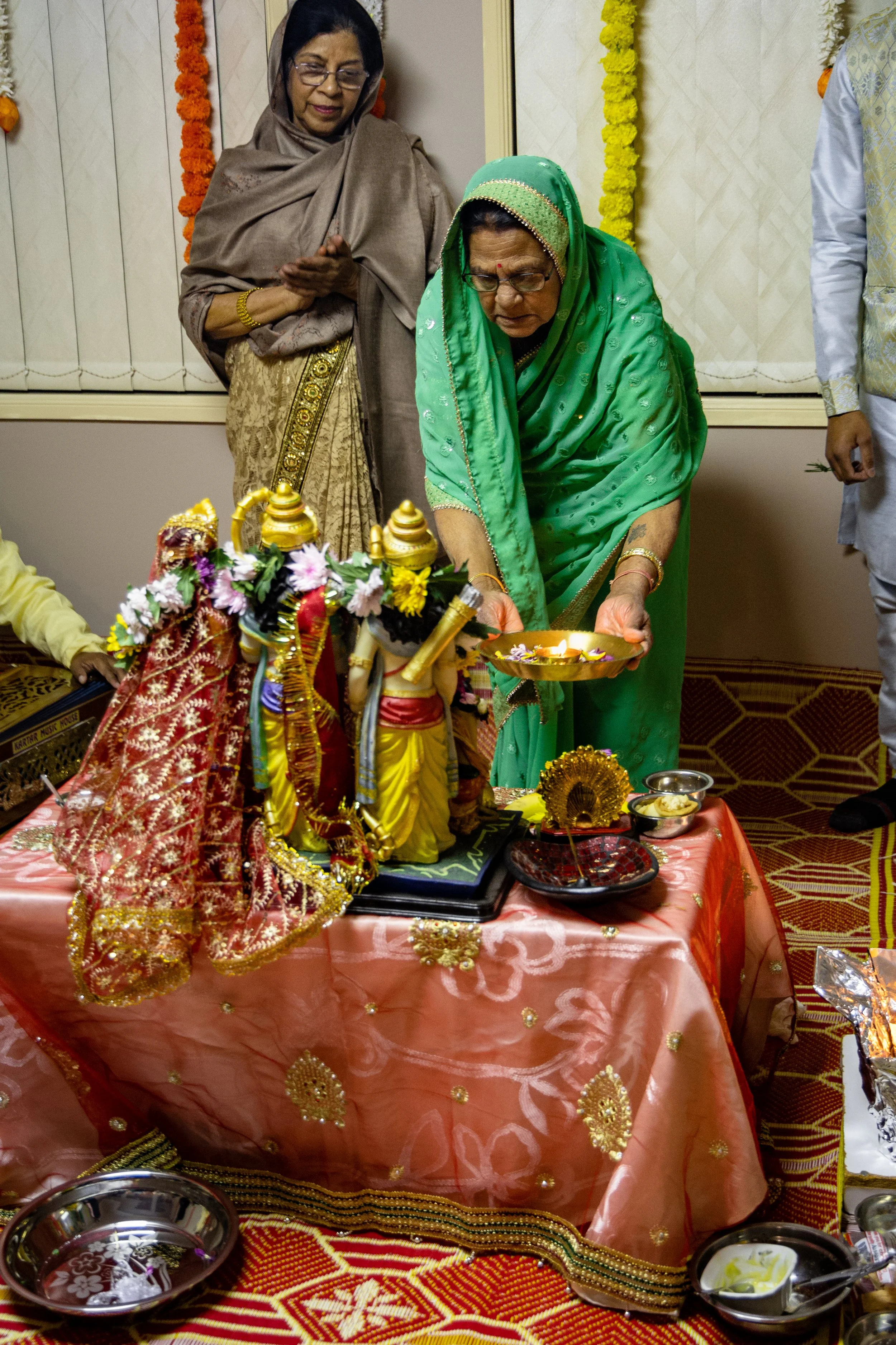 An elderly woman in a green sari and glasses is participating in a religious ceremony, pouring water over a deity idol on a decorated table, with two other women watching nearby.