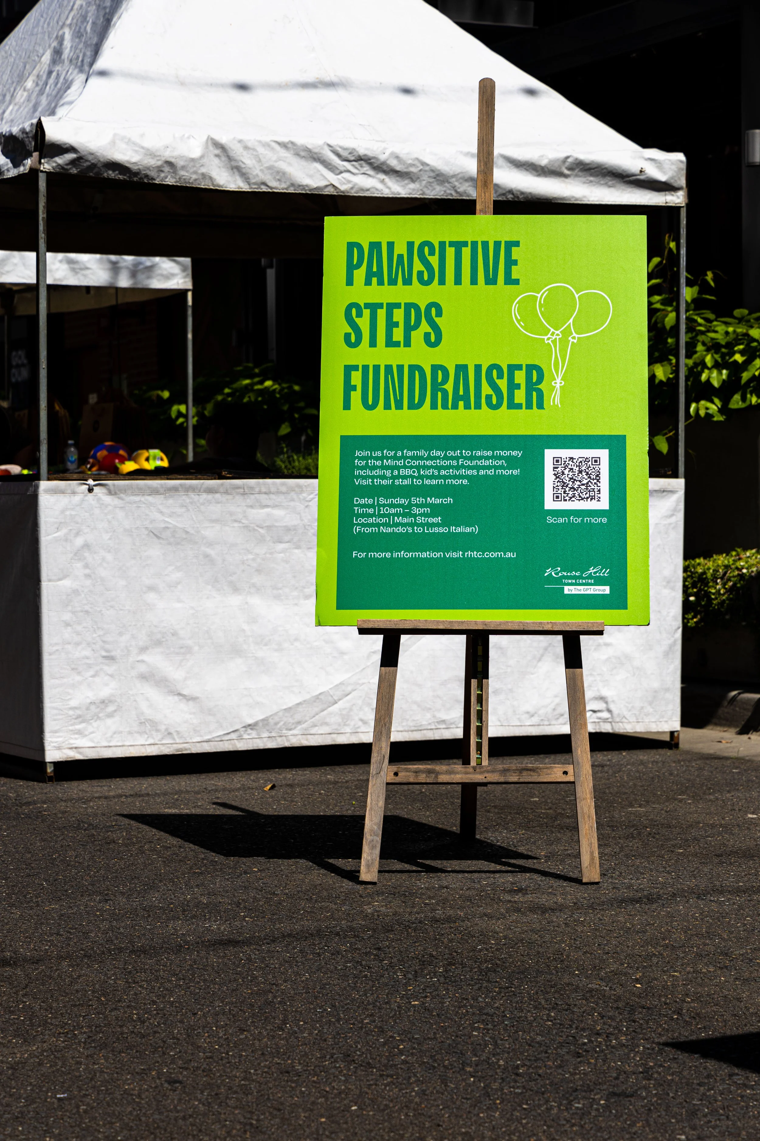 Bright green sign on wooden stand promoting the Pawsitive Steps Fundraiser event, with details about the date, time, location, and a QR code for more information.