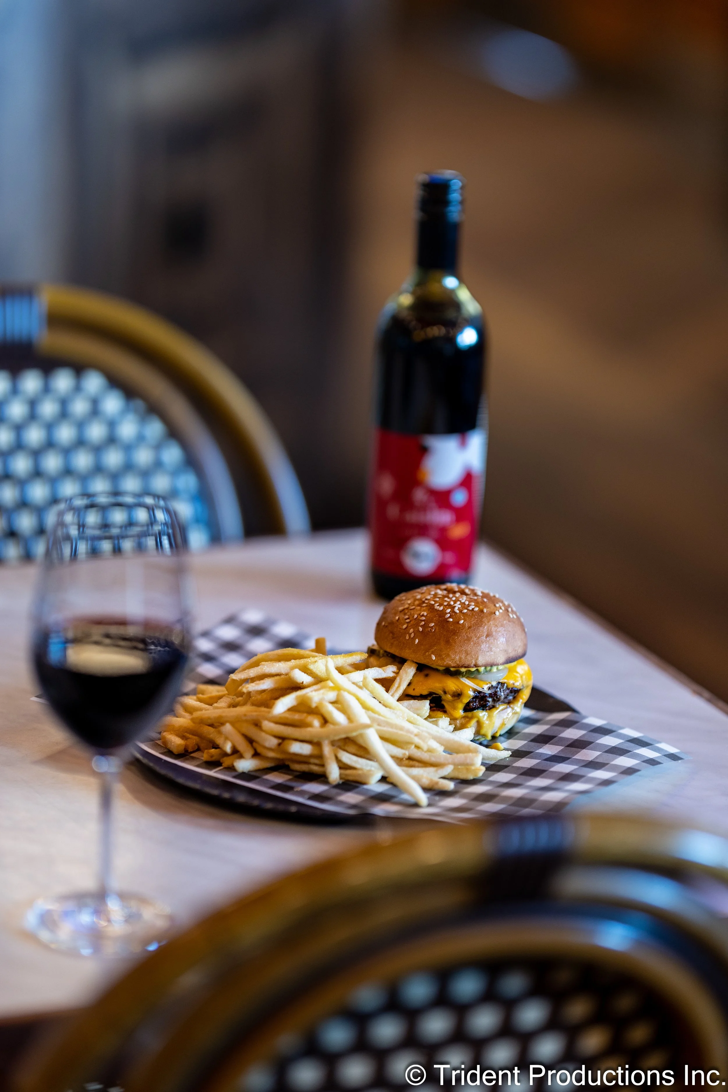 A plate with a cheeseburger and French fries, a glass of red wine, and a bottle of soda on a table with checkered paper, with chairs and a wooden wall in the background.
