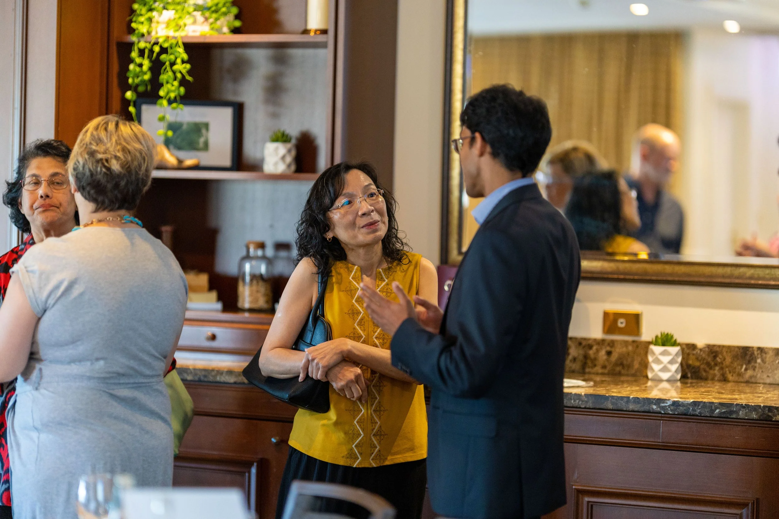 A group of people engaging in conversation at a social event in a warmly decorated room with a wooden bookshelf, plants, and a large mirror.