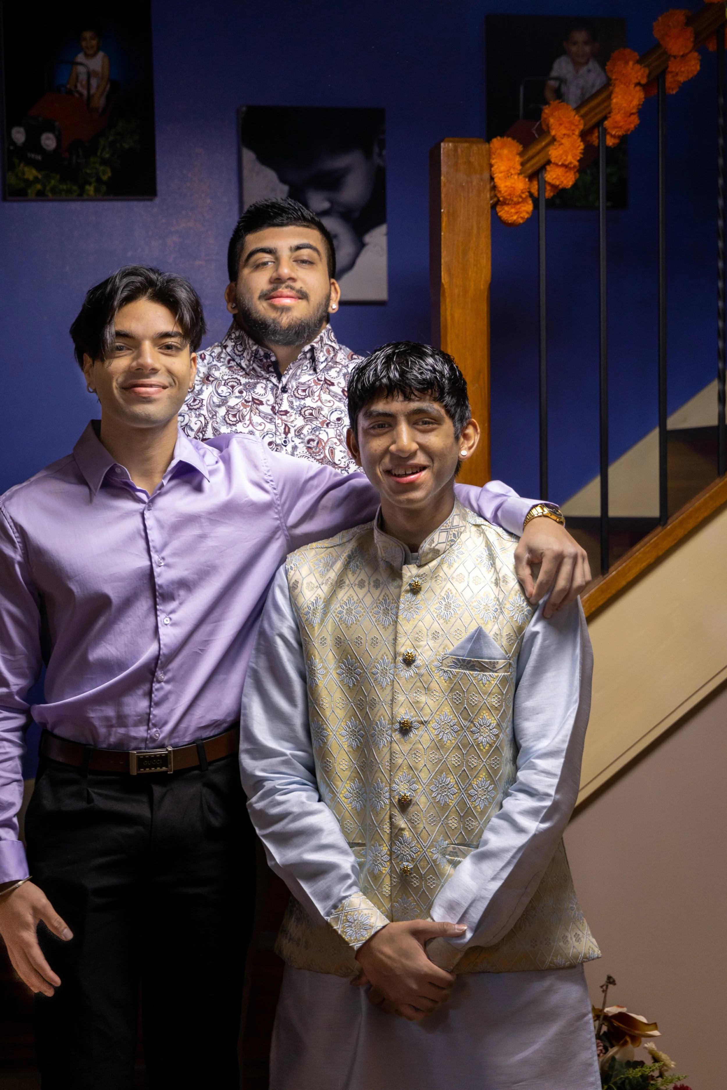 Three young men standing together indoors, one in purple shirt, one in cream-colored traditional Indian attire, and one in patterned shirt, smiling, with a staircase and framed photographs on a blue wall background.