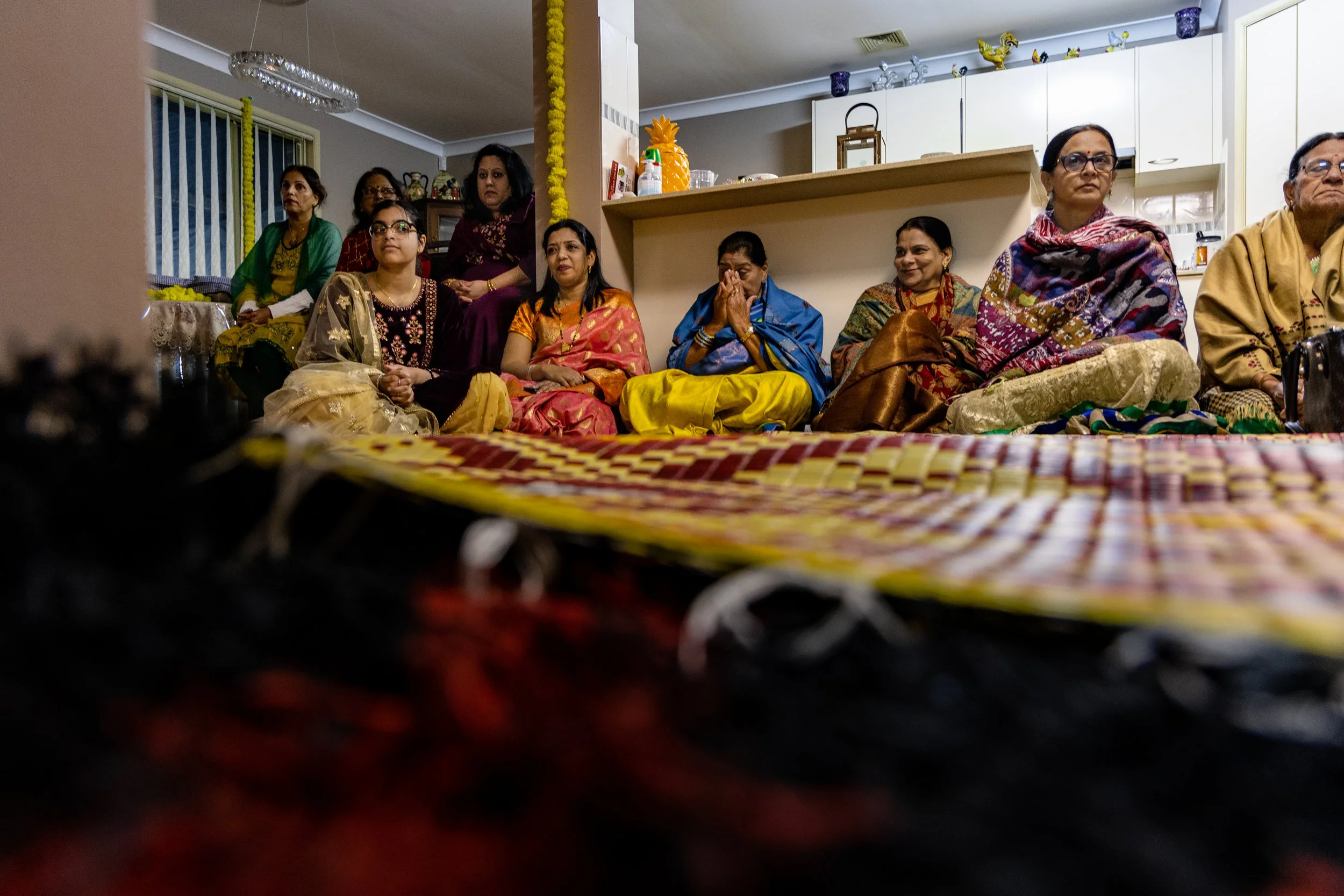 A group of women and girls sitting on the floor in a living room, dressed in traditional Indian clothing, during a celebration or gathering.