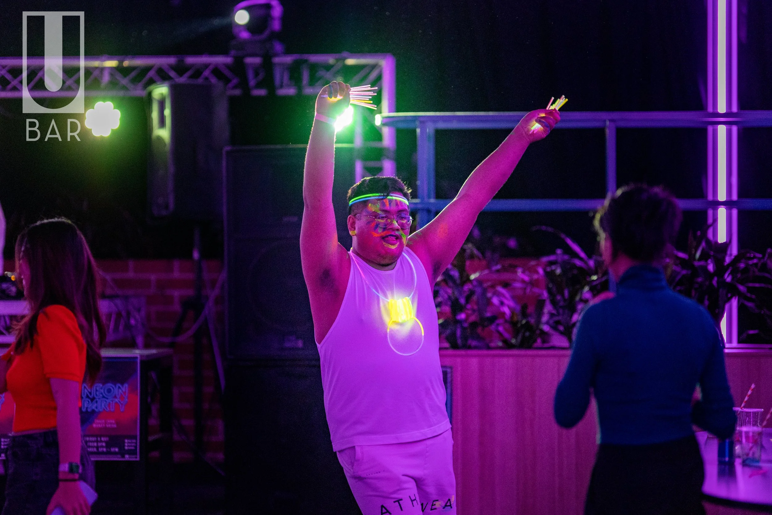 Person dancing at a neon-themed party wearing glow accessories, surrounded by colorful purple and green lighting, with others socializing nearby.