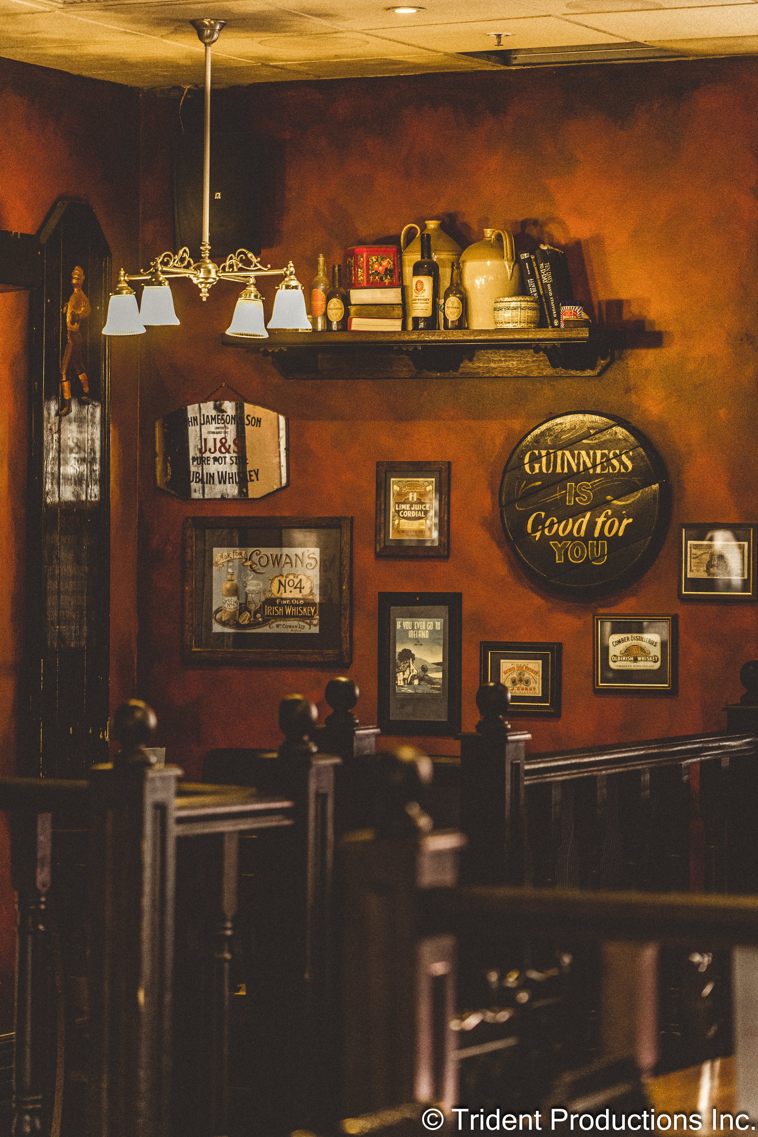 Interior of a cozy pub with dark wooden furniture, vintage wall decor, and a warm, amber light. The wall features framed pictures, a Guinness sign, and a shelf with bottles and books.