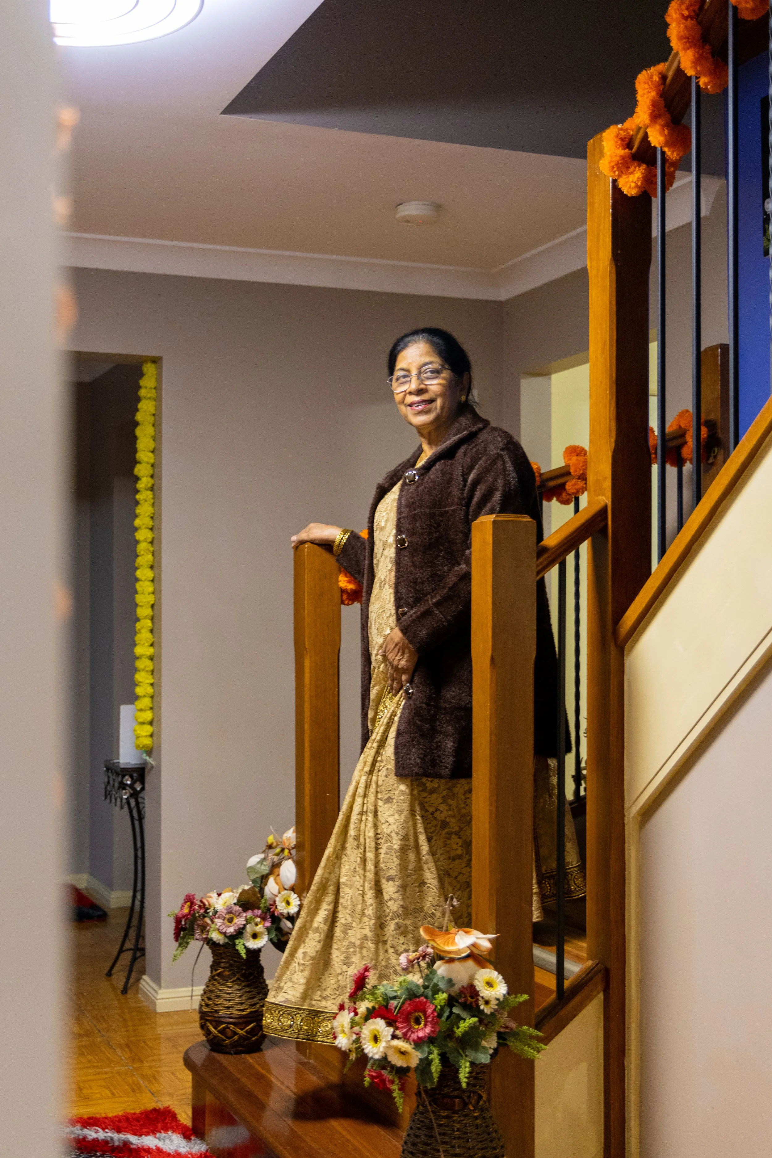 An elderly woman wearing a golden saree and brown coat, smiling while standing on a stairway decorated with orange marigold flowers, in a home with floral arrangements on the floor.