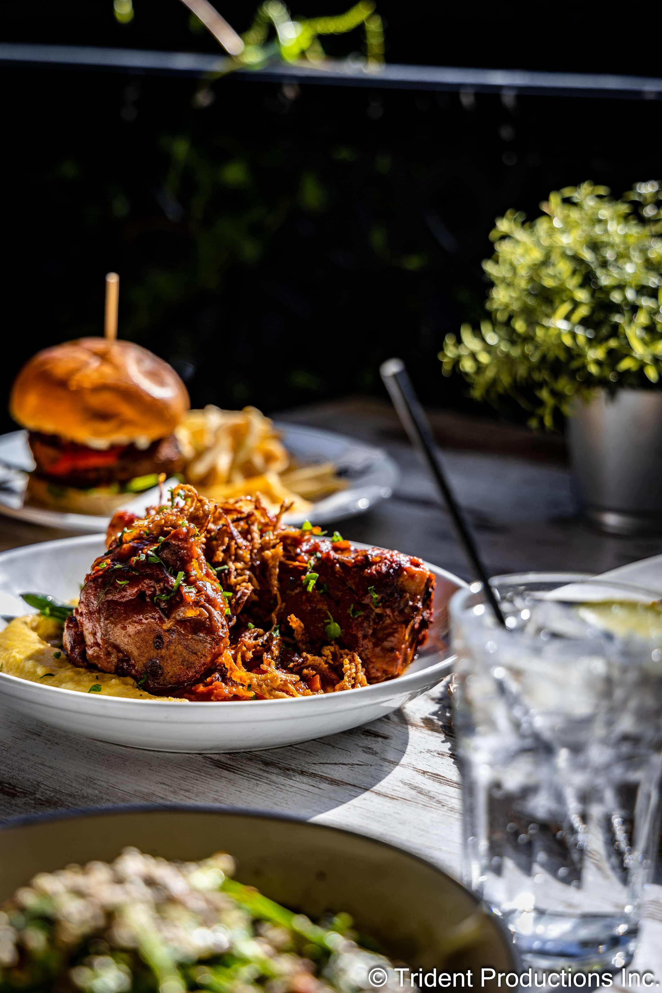 Close-up of a plate with braised meat, mashed potatoes, and herbs, with other plates of food, a drink, and a burger in the background on a wooden outdoor table.