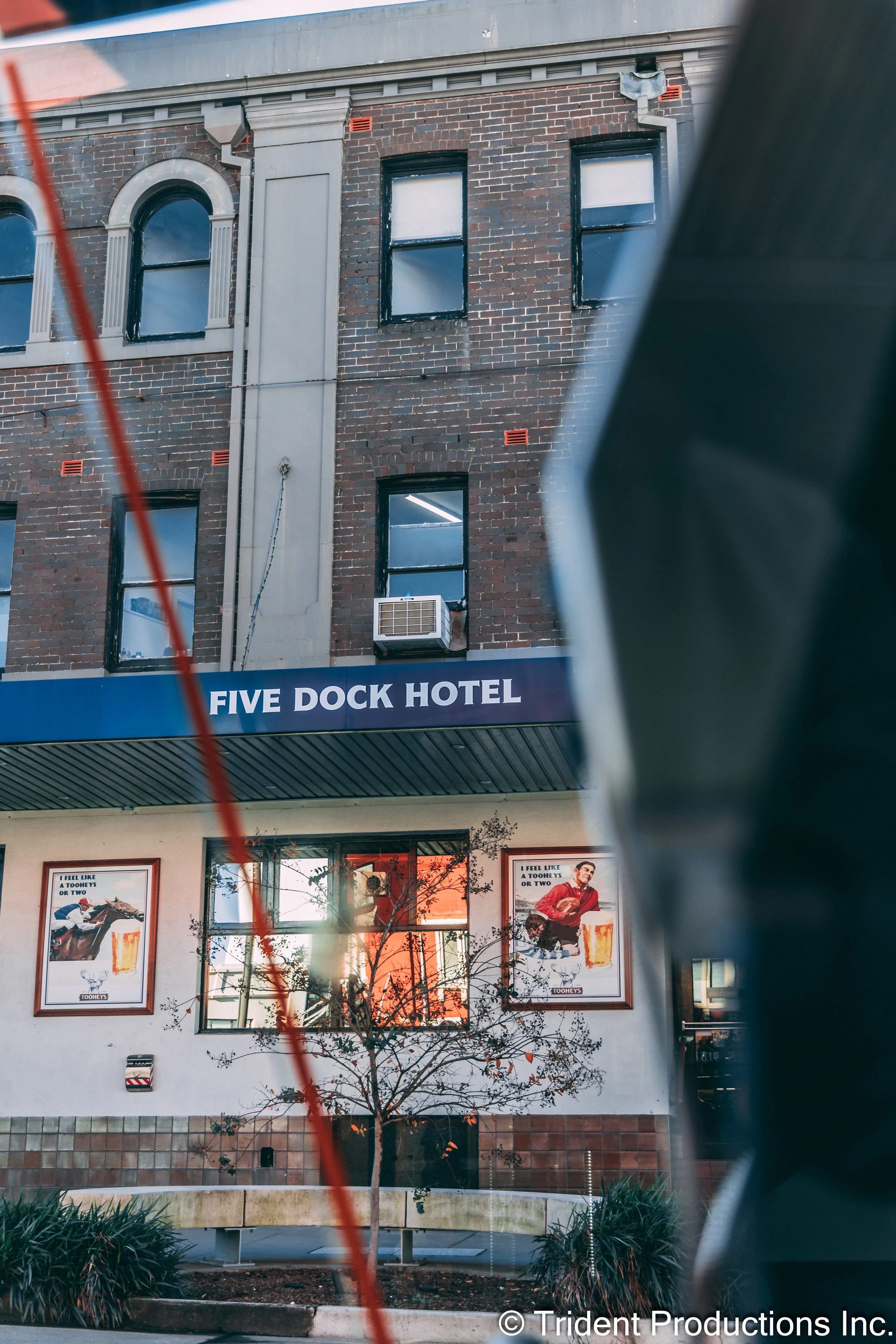 Exterior of Five Dock Hotel with brick building facade, windows, an air conditioning unit, and posters on the wall, with a tree and plants in the foreground.