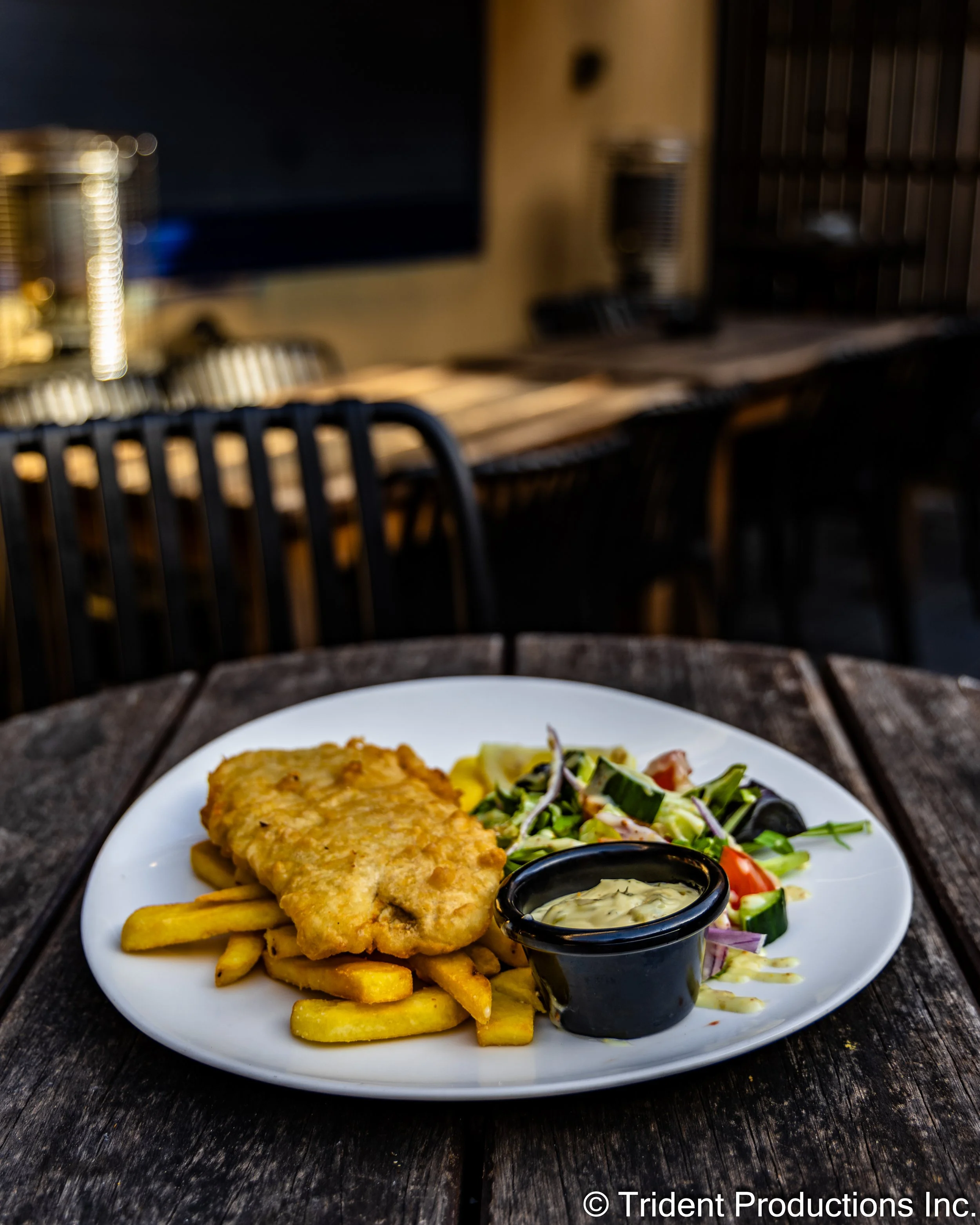 Plate with fried fish fillet, French fries, salad, and dipping sauce on a wooden table in a dimly lit restaurant.