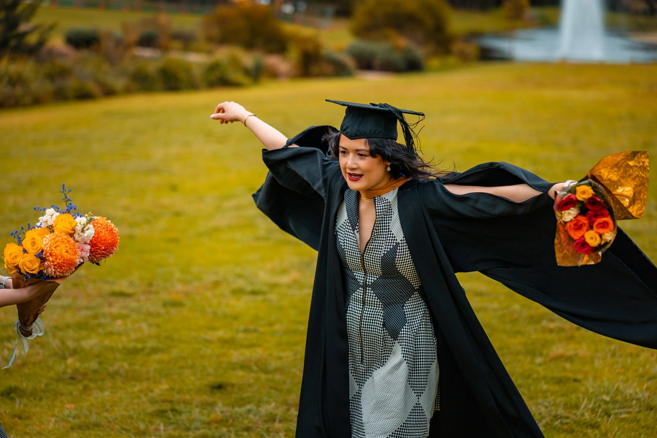A young woman in a graduation cap and gown celebrating outdoors on a grassy area, holding a bouquet of colorful flowers in each hand, with a fountain in the background.