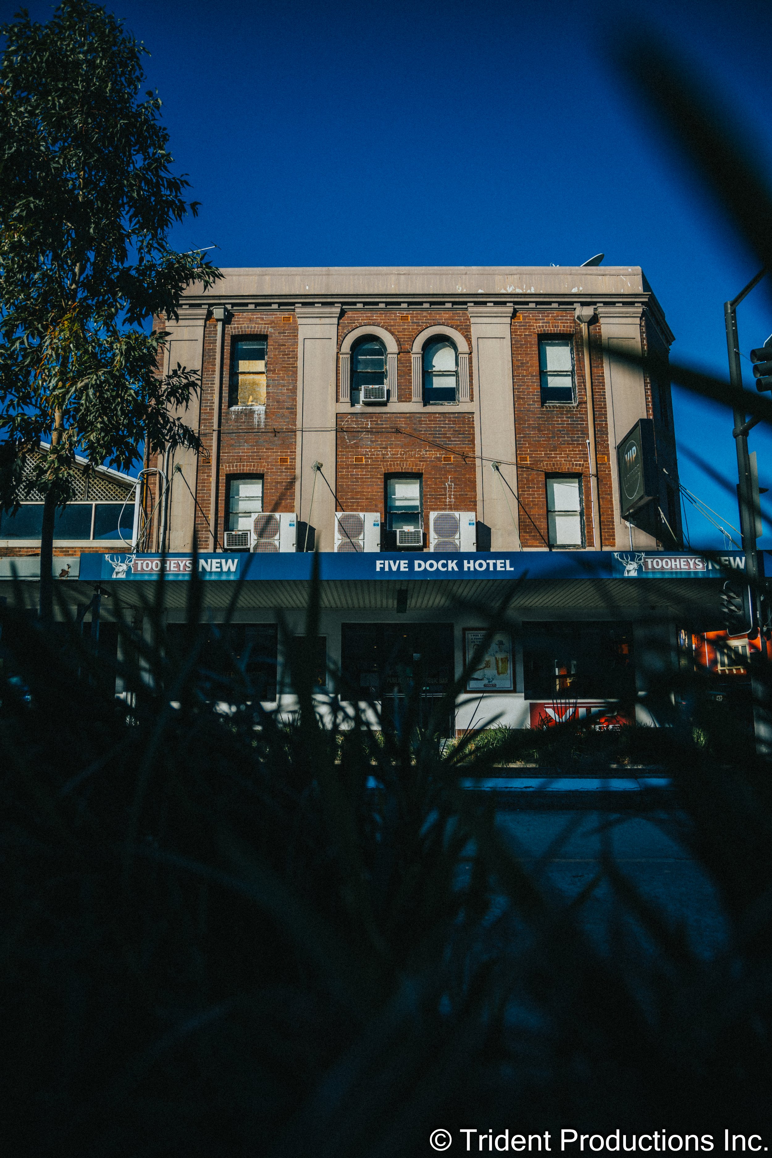 A brick building with a sign that reads 'Five Dock Hotel' and air conditioning units on the front. The building has ornamented window arches and a tree on the left, with a clear blue sky above.