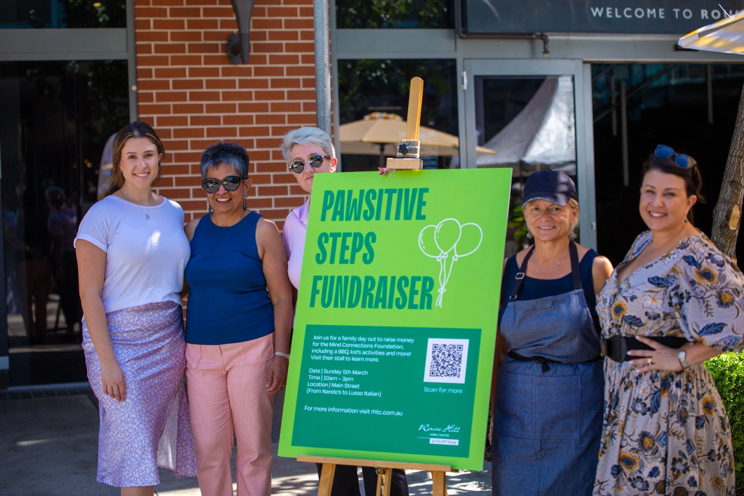 Group of five women standing outdoors next to a green sign for the Pawsitive Steps Fundraiser, with a brick building and glass doors in the background.