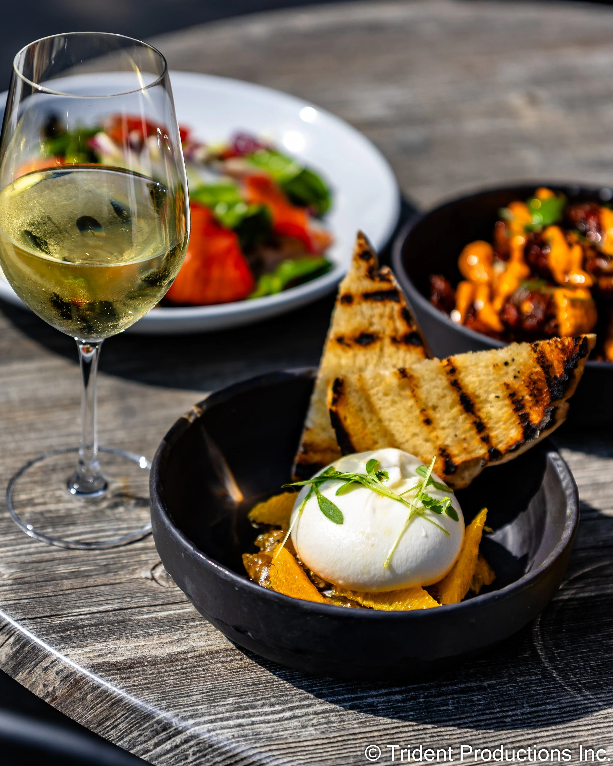 A glass of white wine, a small bowl of roasted vegetables with creamy topping, grilled bread slices, and a side salad with various vegetables on a wooden table.