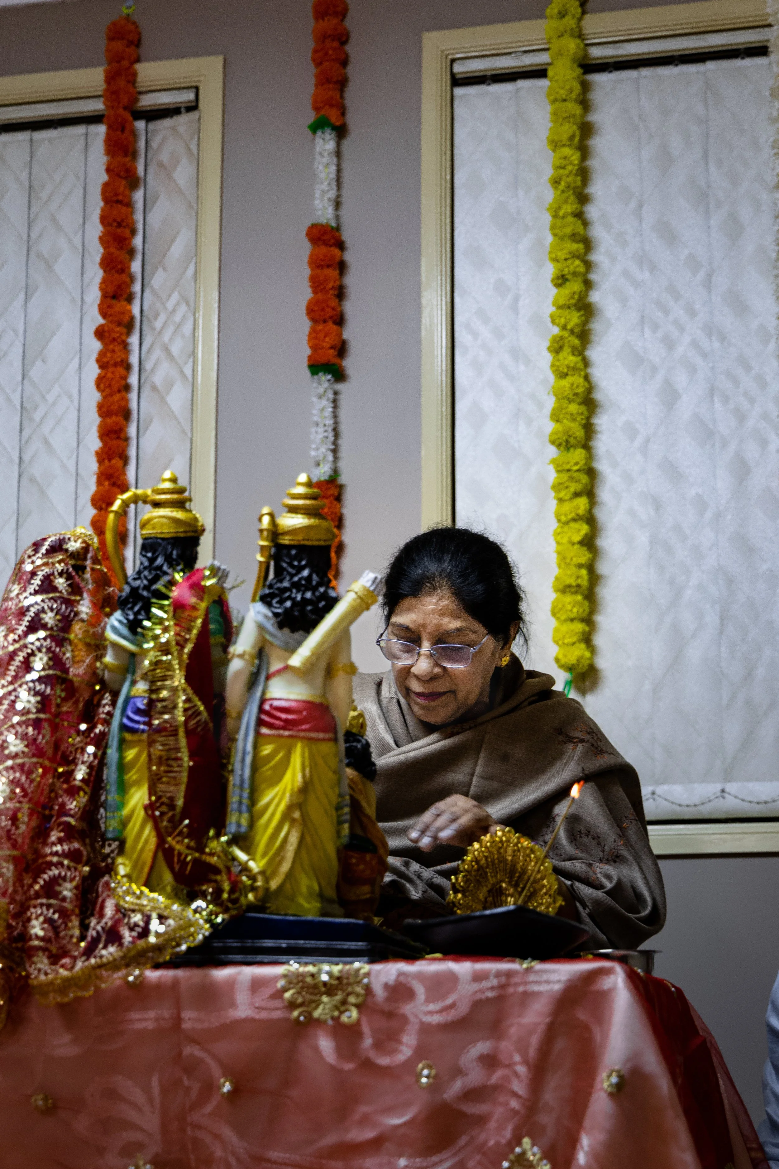 An elderly woman with glasses is participating in a traditional Hindu ritual, placing offerings near idols of Lord Rama, Sita, Lakshmana, and Hanuman, decorated with ornate clothing and jewelry, on a table covered with a pink cloth.