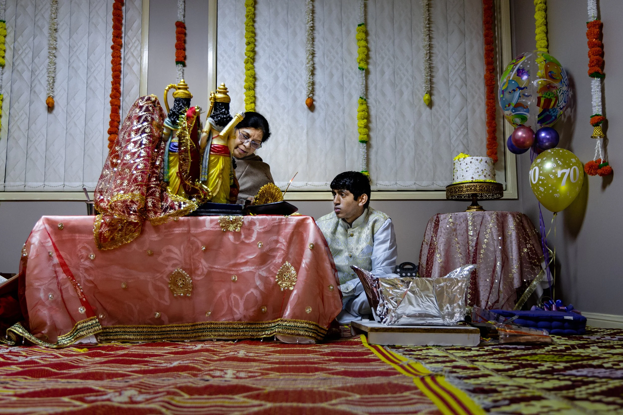 An Indian family participating in a religious ceremony or ritual at home, with decorations including balloons, flowers, and a decorated table with a religious idol.