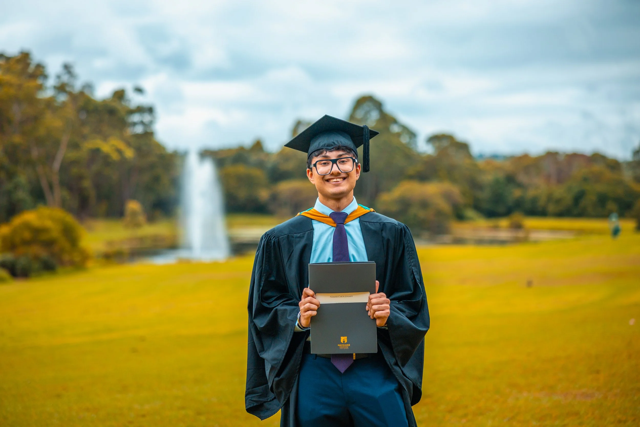 Graduate in cap and gown holding diploma outdoors with a fountain and trees in the background.
