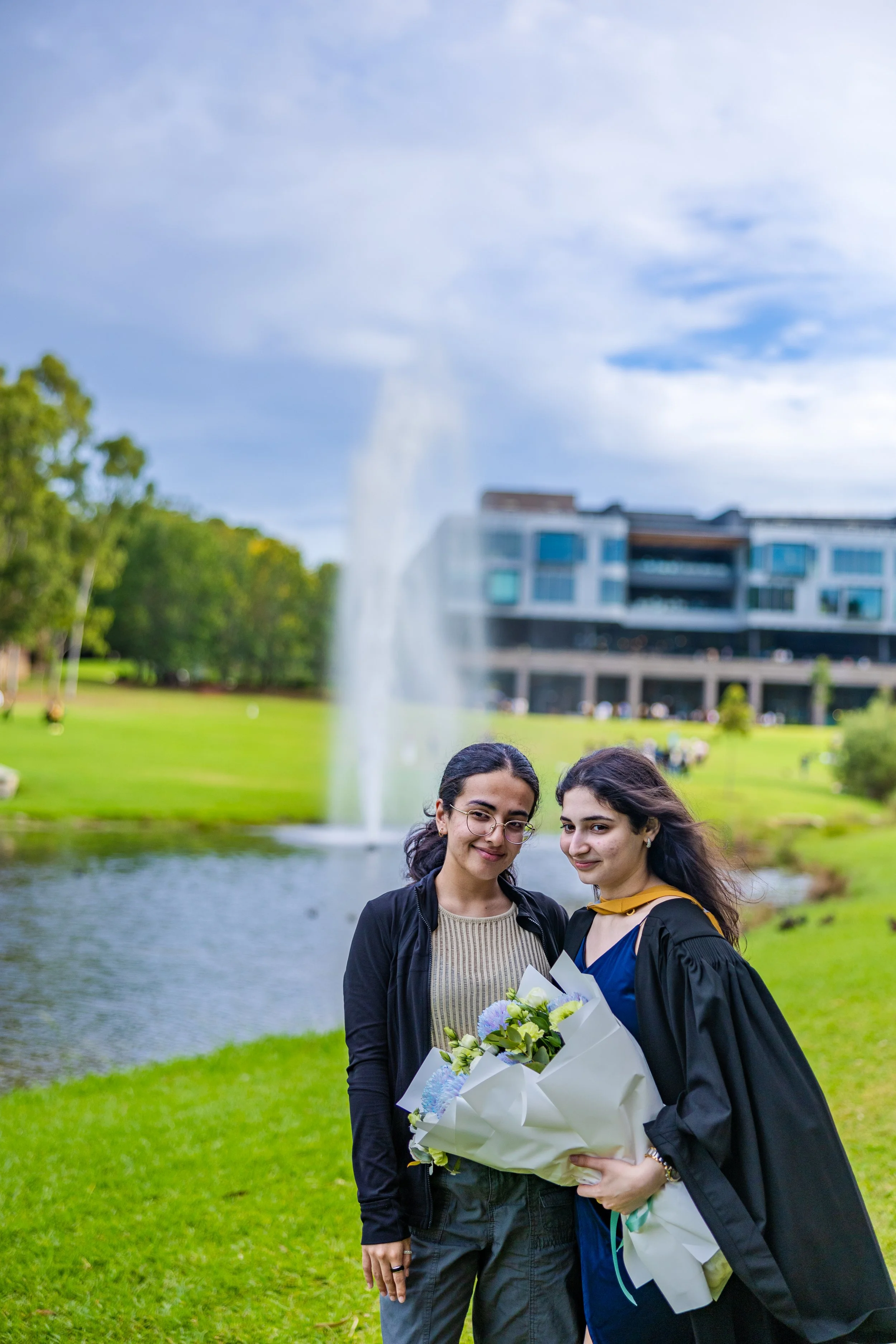 Two women standing outdoors near a pond with a fountain, one wearing a graduation gown holding flowers, and the other in casual clothing, posing for a photo during a graduation celebration.