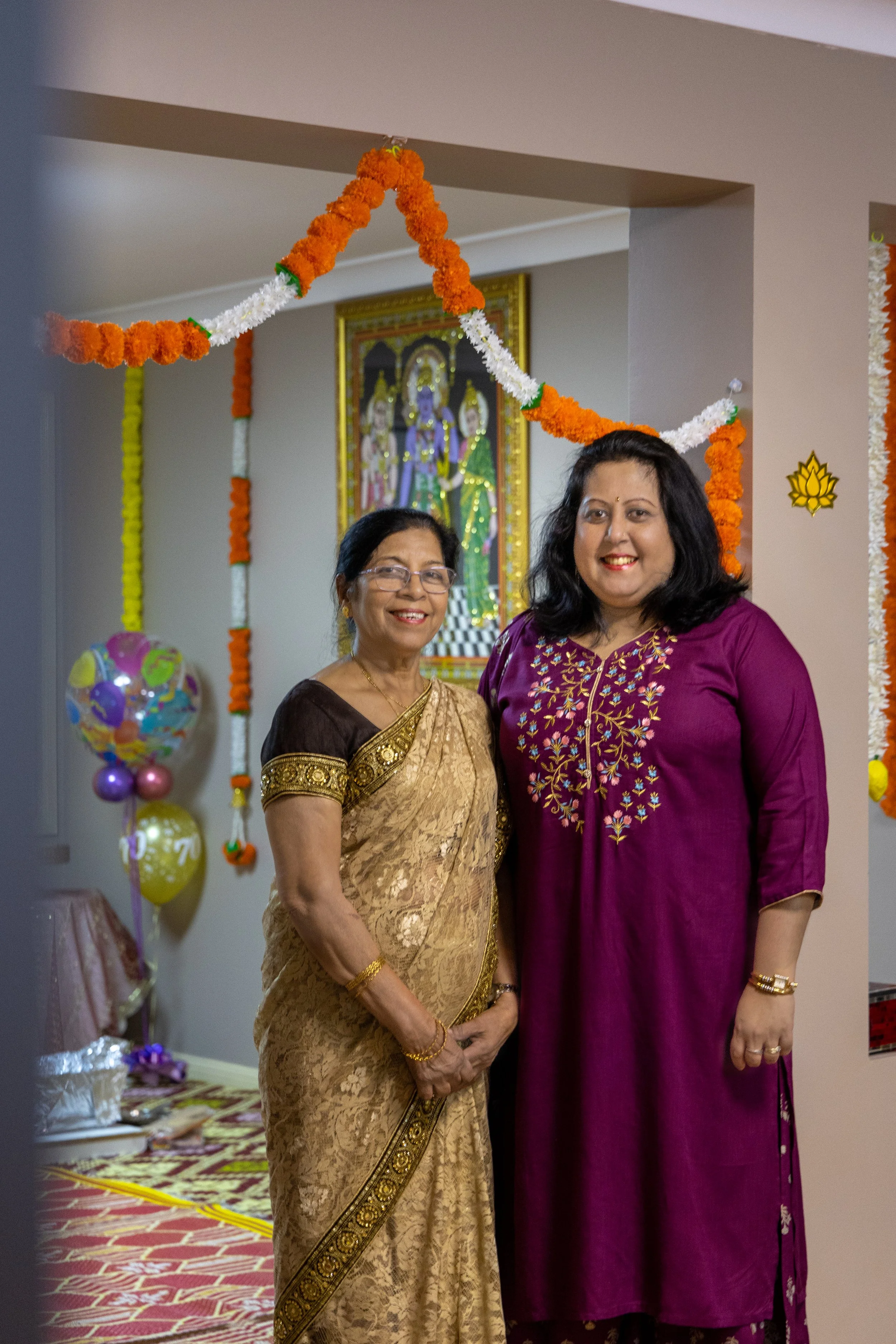 Two women dressed in traditional Indian saree and kurta smiling at the camera, standing in a decorated room with Hindu religious images, balloons, and floral garlands.