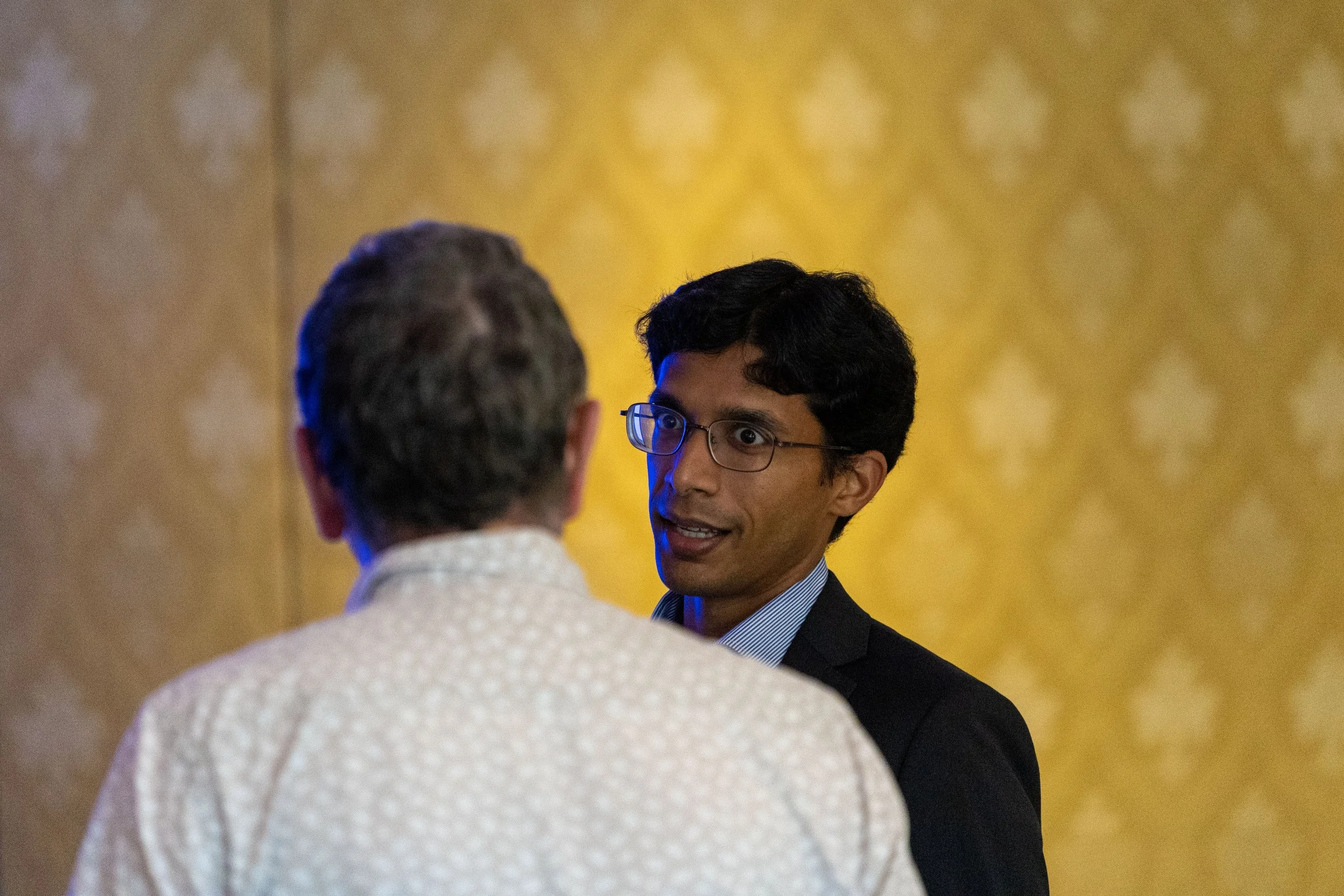 Two men engaged in conversation, one facing the camera with glasses and dark hair, and the other with gray hair seen from behind, in a room with yellow patterned wallpaper.