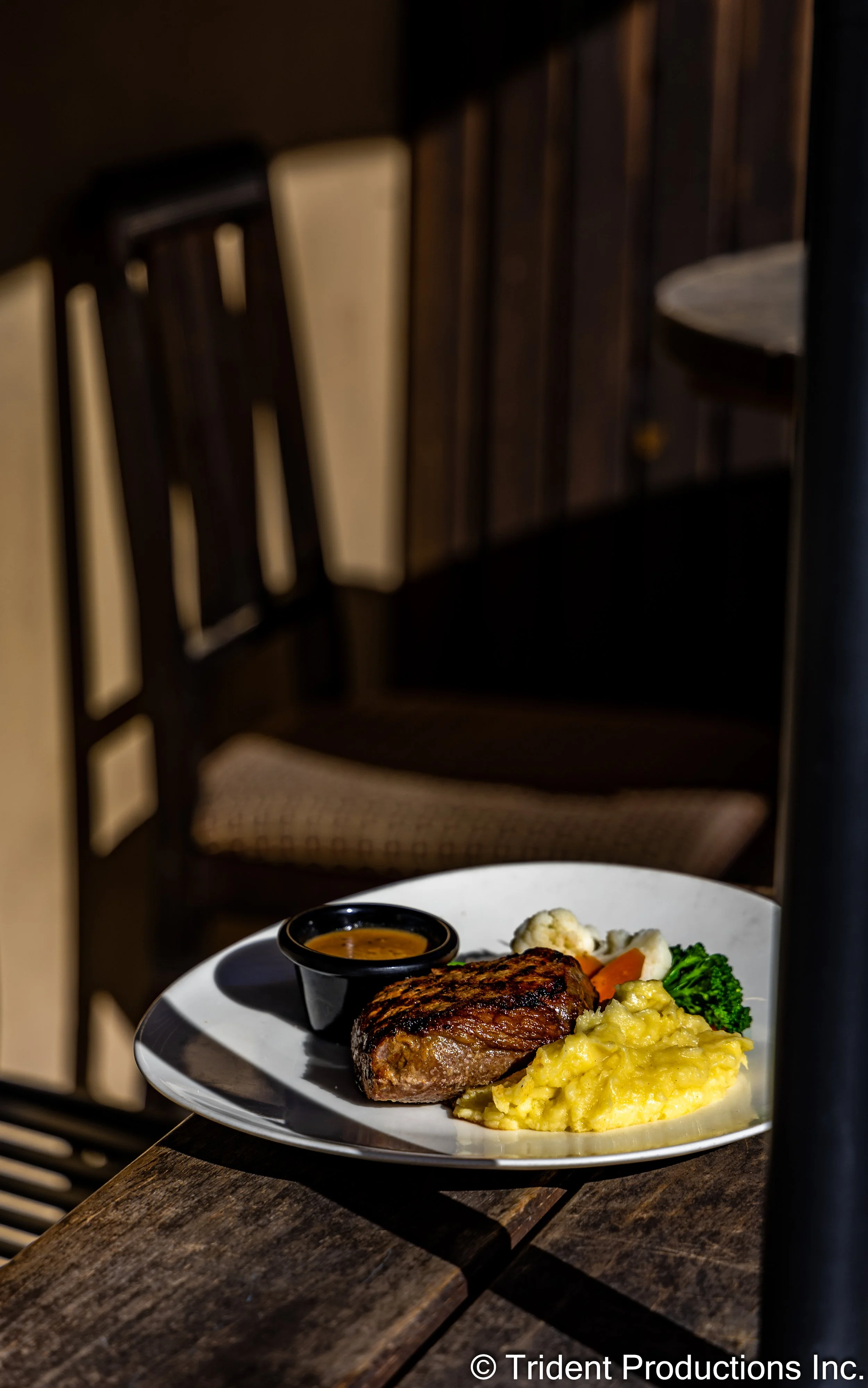 Plate with steak, mashed potatoes, broccoli, cauliflower, and a small cup of gravy or sauce, on a wooden table.