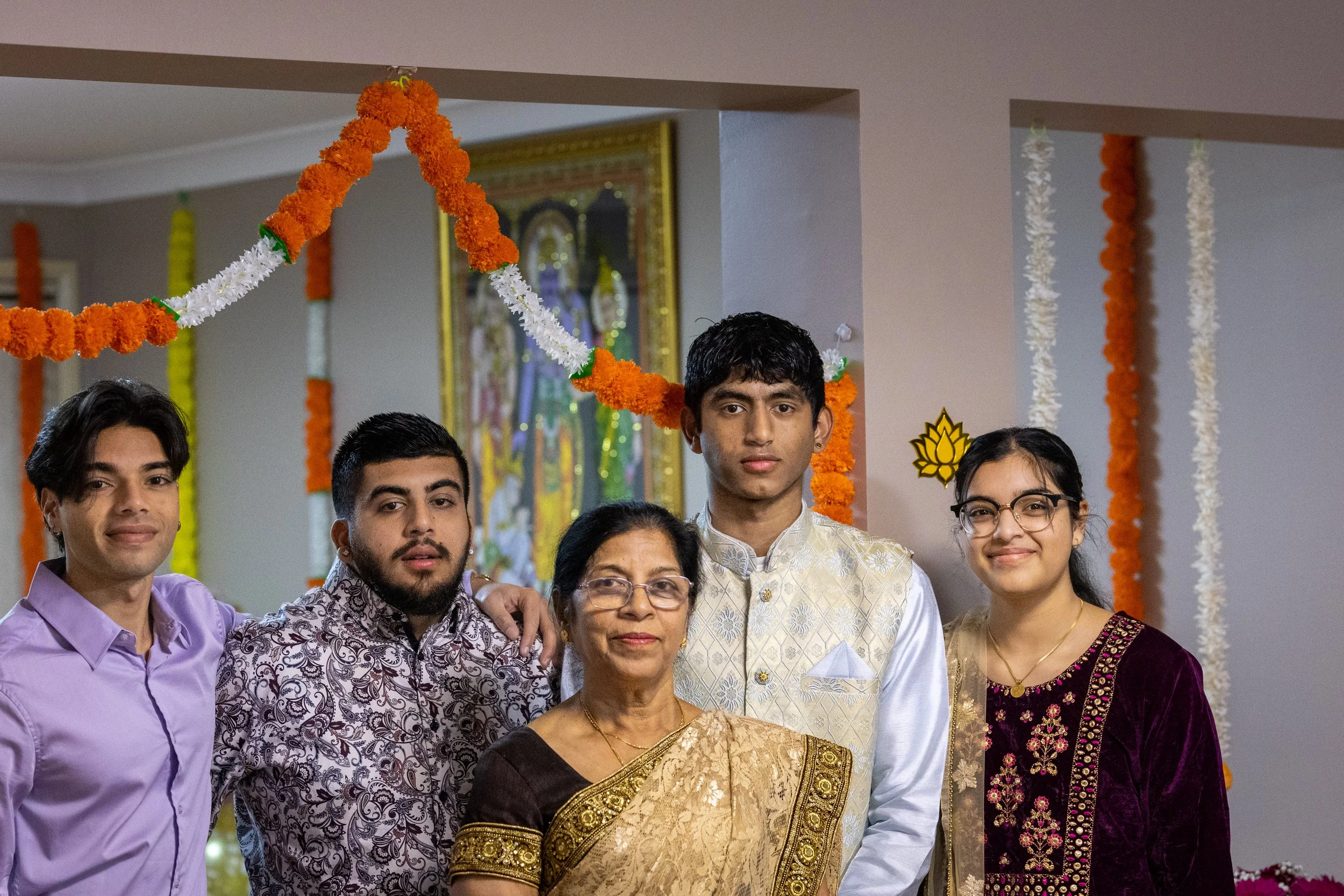 Family of five celebrating a traditional Indian festival, with colorful flower garlands and religious artwork in the background.