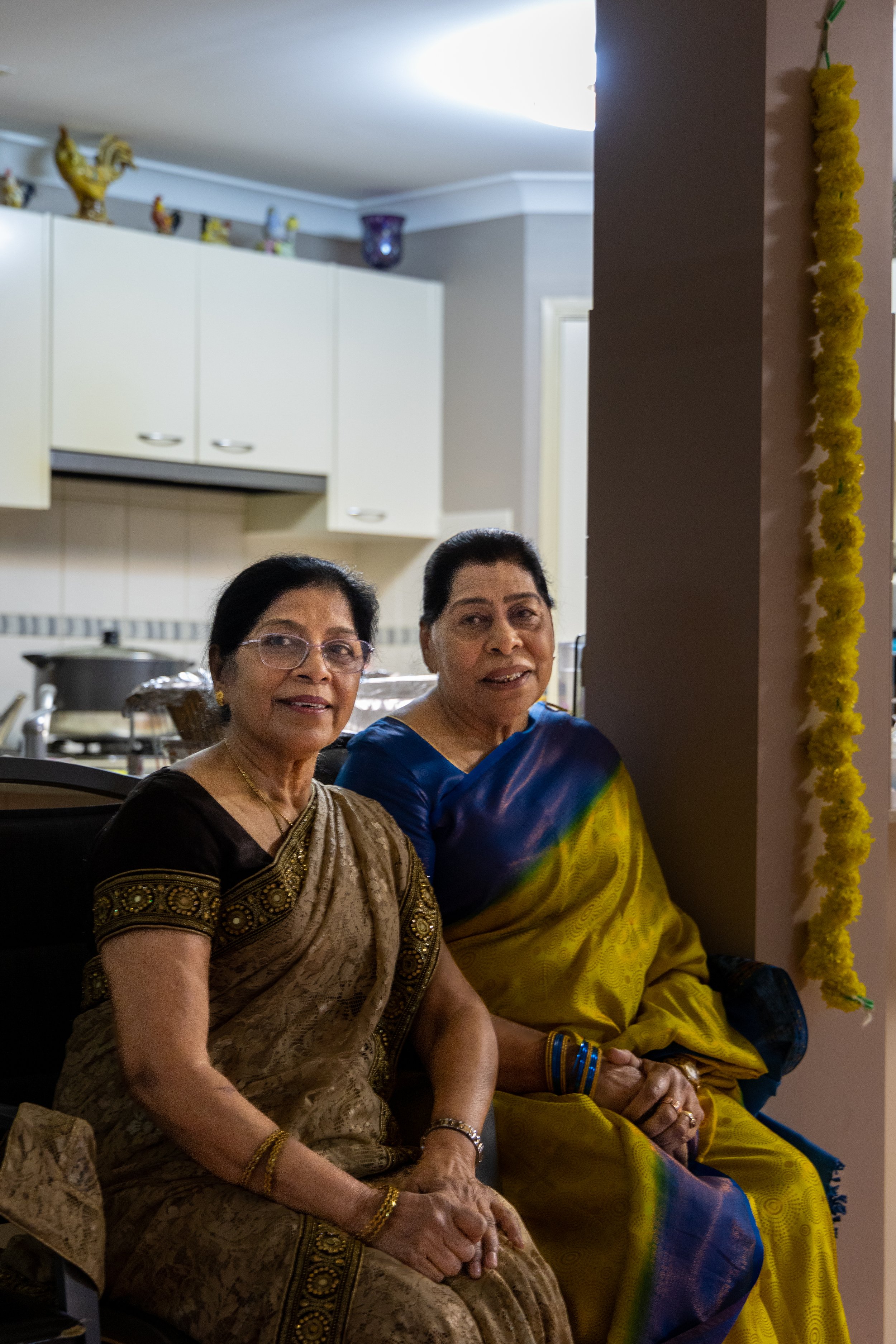 Two elderly women sitting in a living room, dressed in traditional Indian sarees, looking at the camera. One is wearing a brown and gold saree with glasses, the other in a blue and yellow saree.