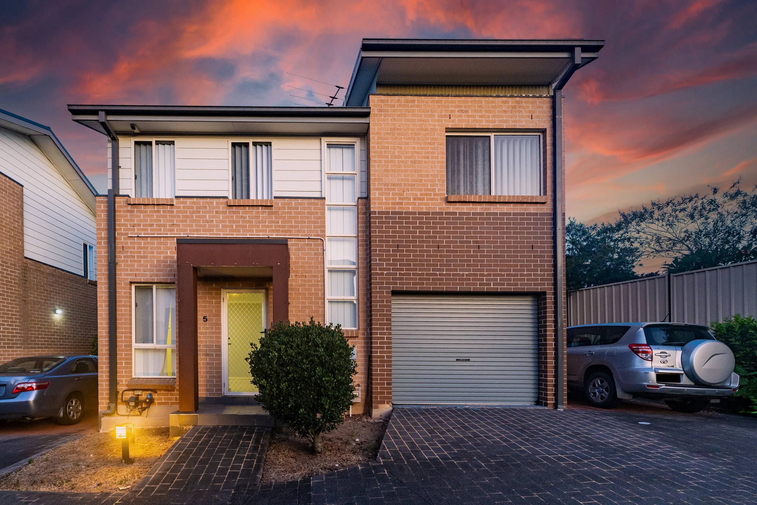 Modern two-story brick and siding house with a garage, surrounded by parked cars, dusk sky with pink and orange clouds.