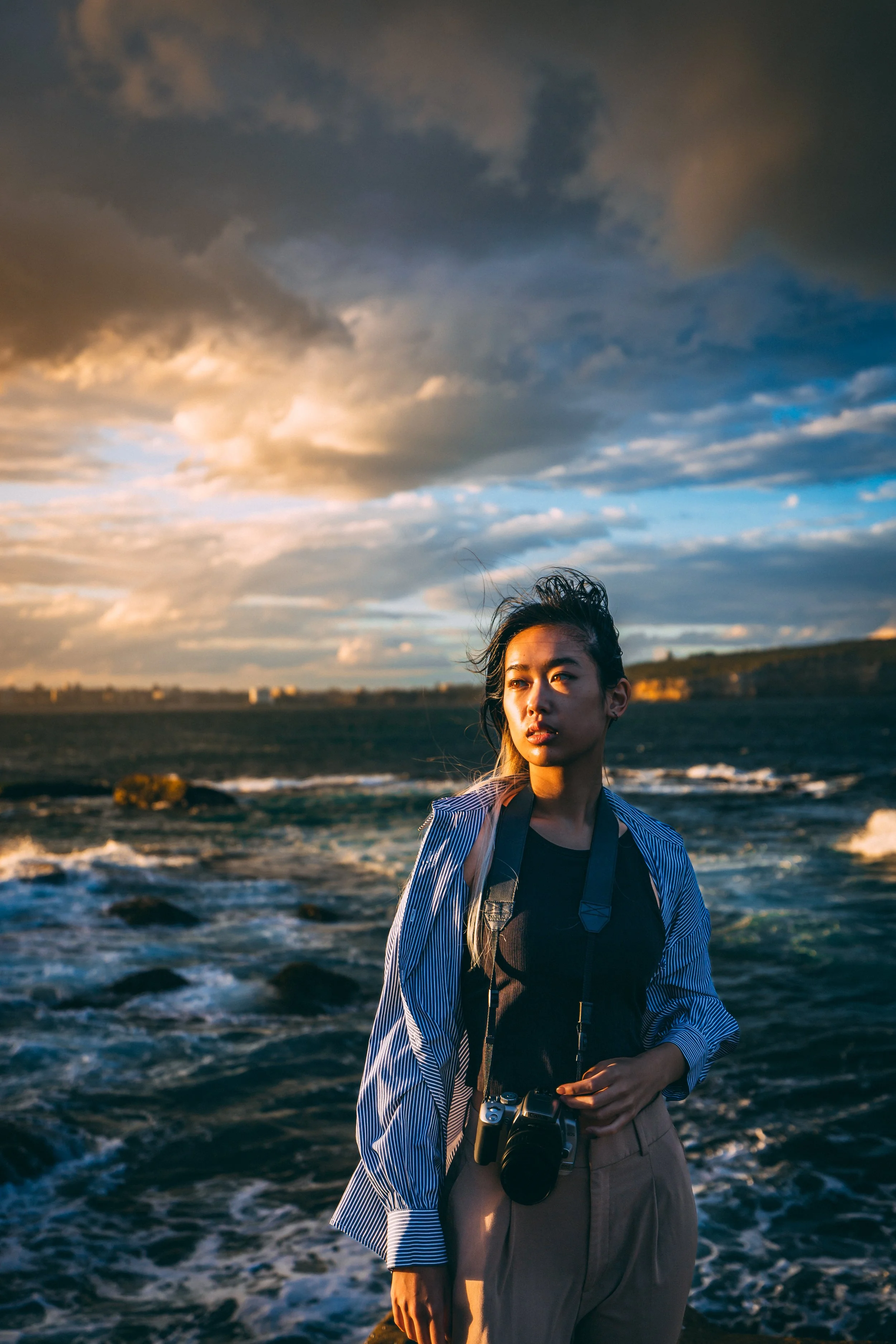 A young woman standing near the ocean during sunset, holding a camera, with clouds and a distant shoreline in the background.
