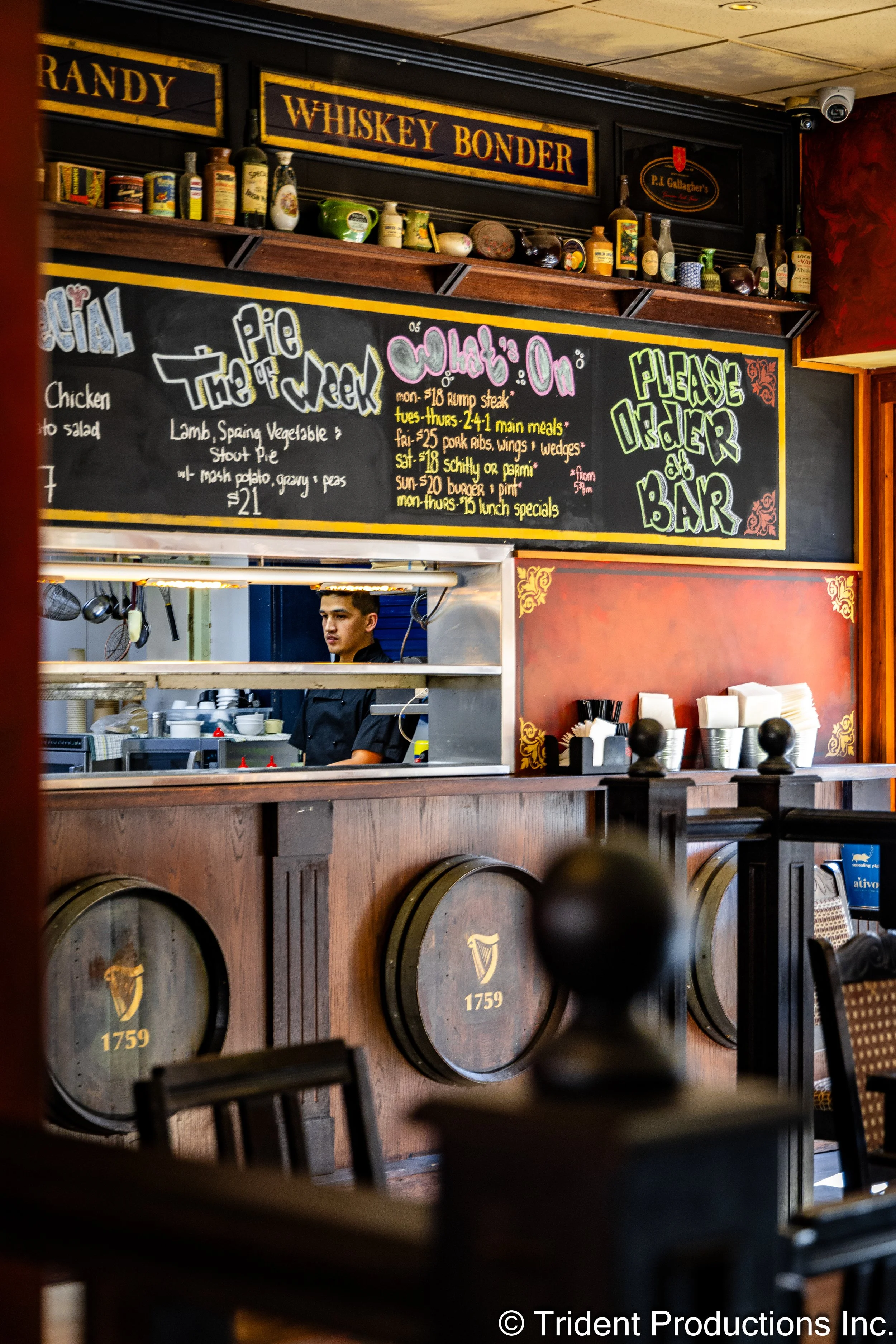Bar area inside a pub with a bartender behind the counter, wooden barrels with Guinness logo, chalkboard menu advertising pie of the week and lunch specials, shelves with alcohol bottles and decor, and a chalkboard sign requesting orders at the bar.