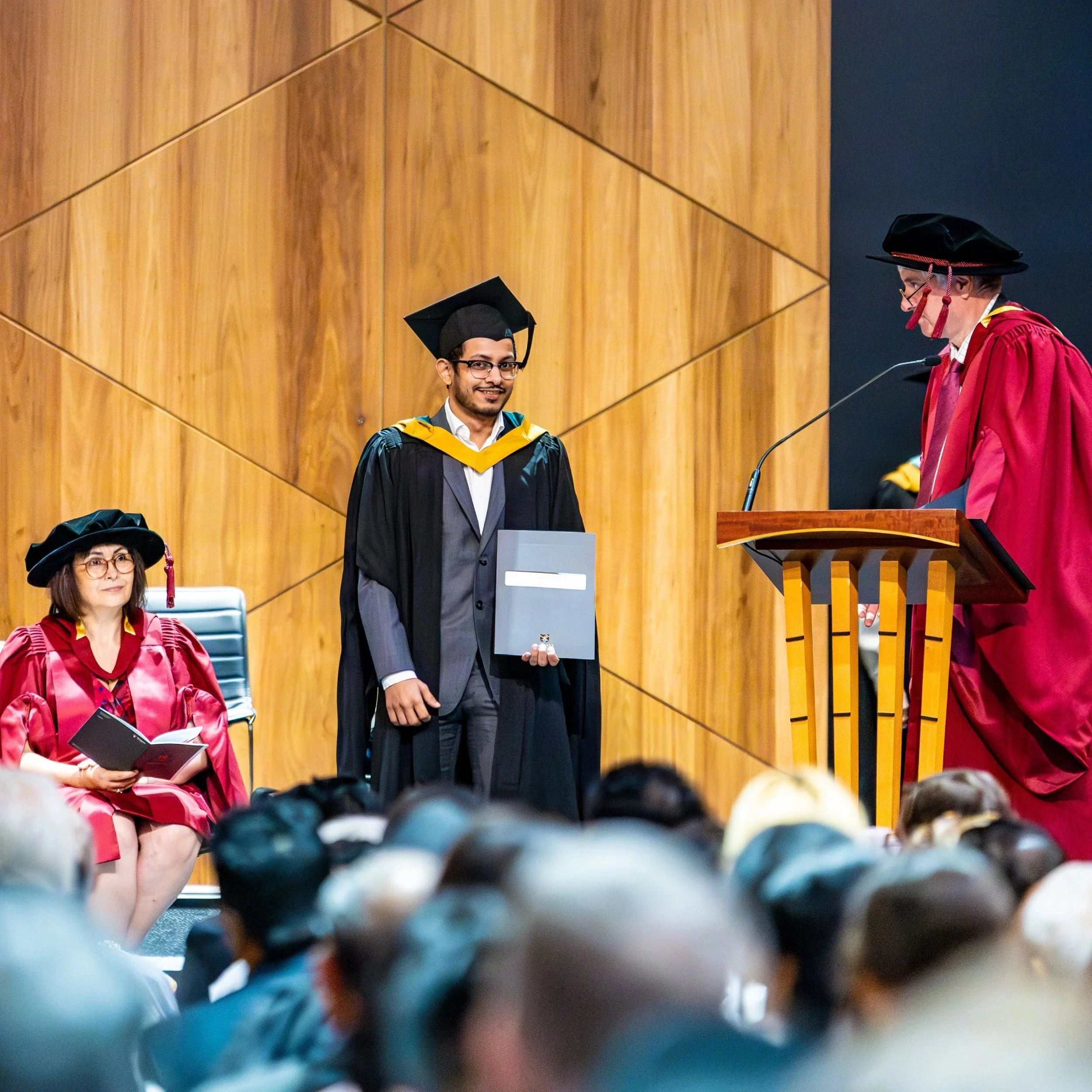 A young man in graduation robes and cap standing on stage receiving diploma during graduation ceremony. Two women in academic regalia seated nearby, and an older man in academic dress speaking at podium.