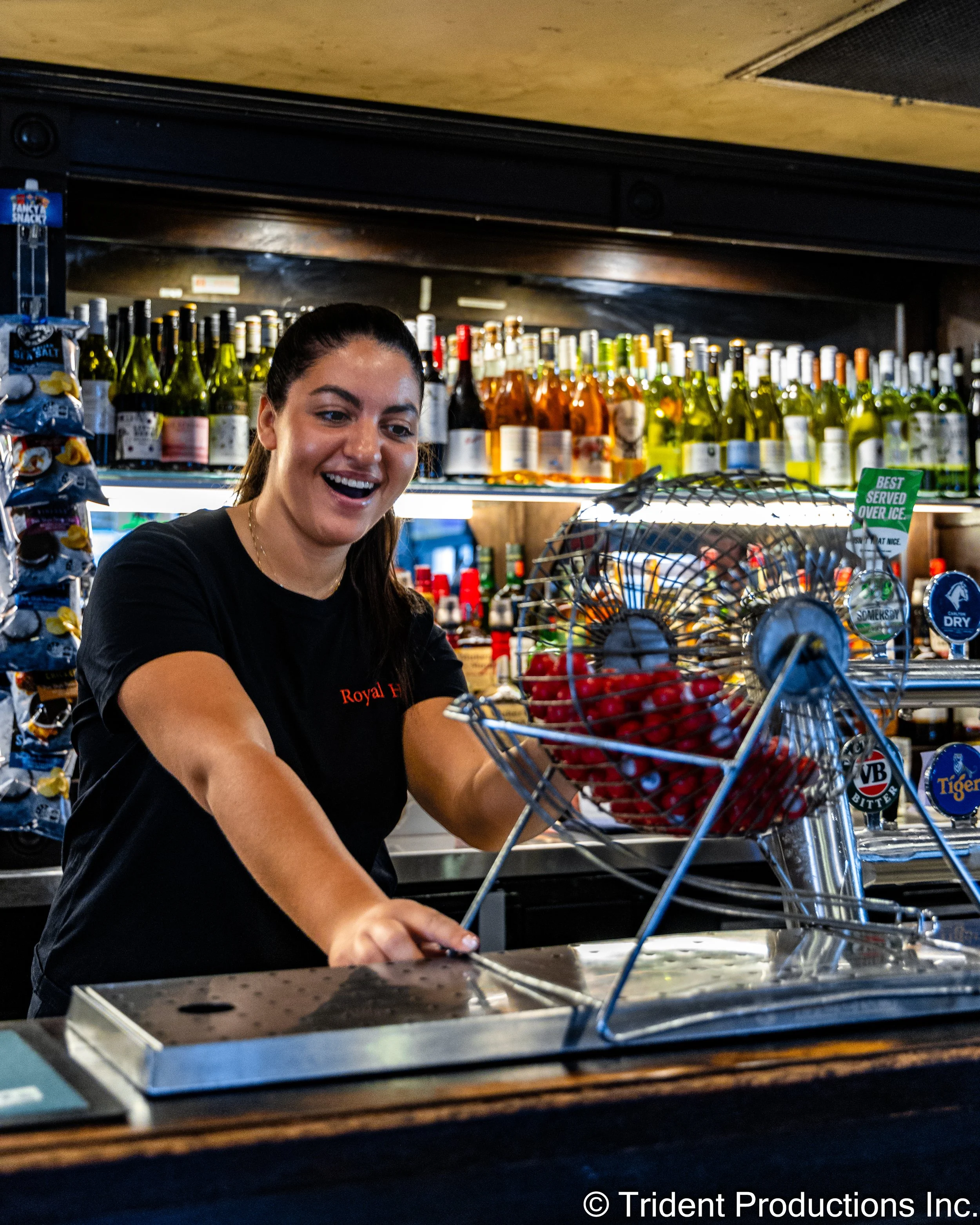 A woman working behind a bar or counter with bottles of wine on shelves in the background and a basket of cherries in front of her.