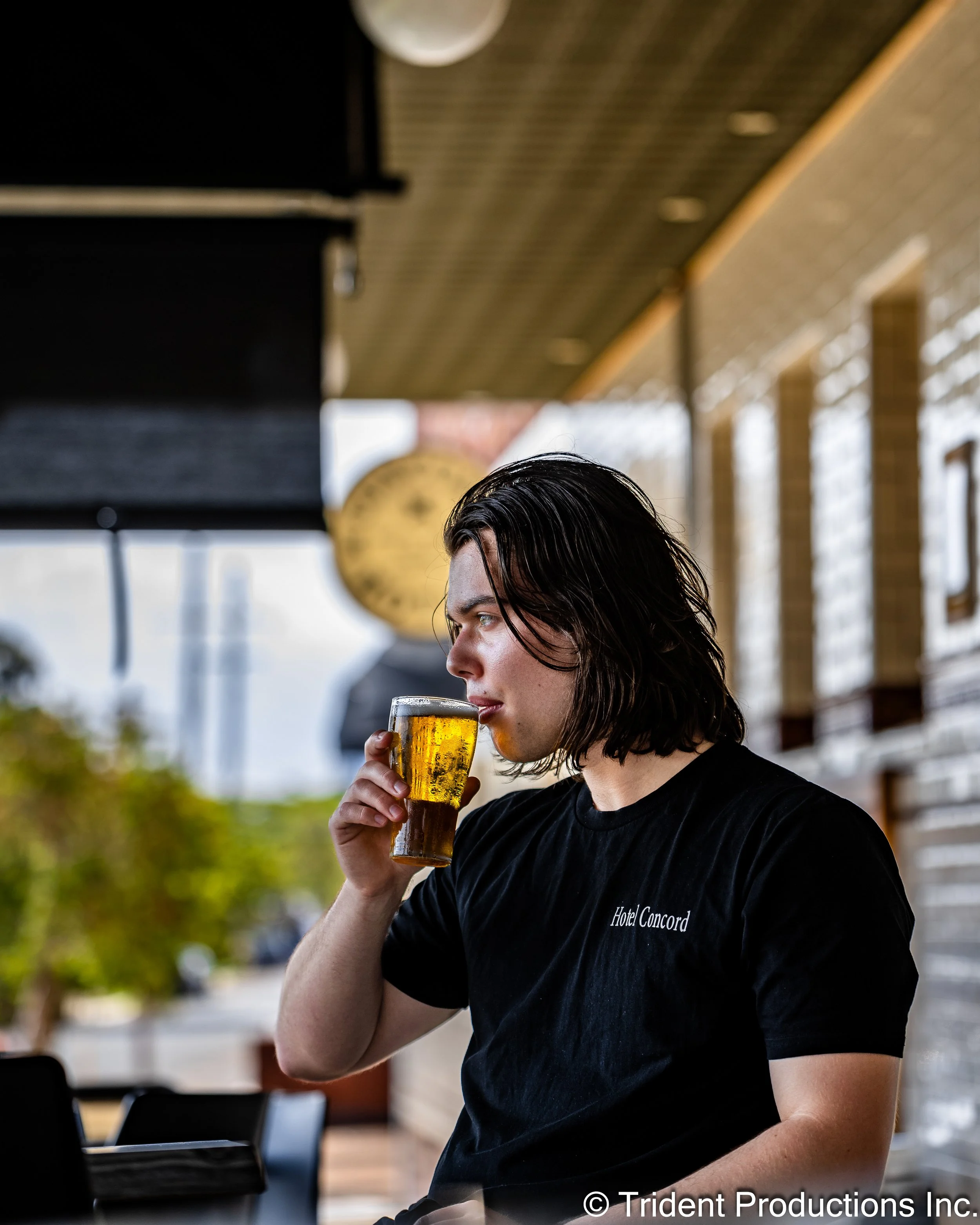 A young man with shoulder-length dark hair is drinking a glass of beer on an outdoor patio at a restaurant or bar.