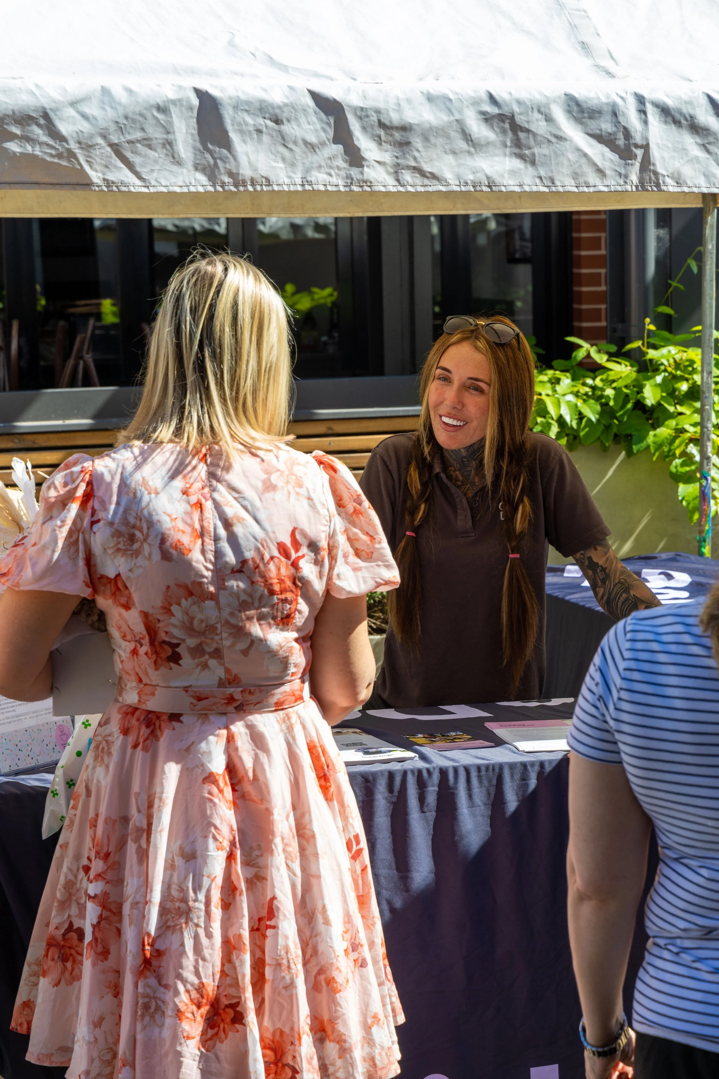 A woman with long red hair, tattoos, and sunglasses on her head is smiling while talking to a woman in a floral dress at an outdoor booth during daytime.