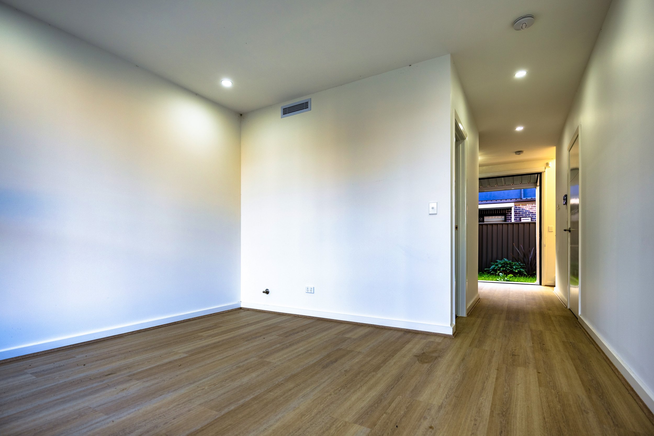 Empty room with white walls, wooden flooring, and a hallway leading outside with a fence and garden.
