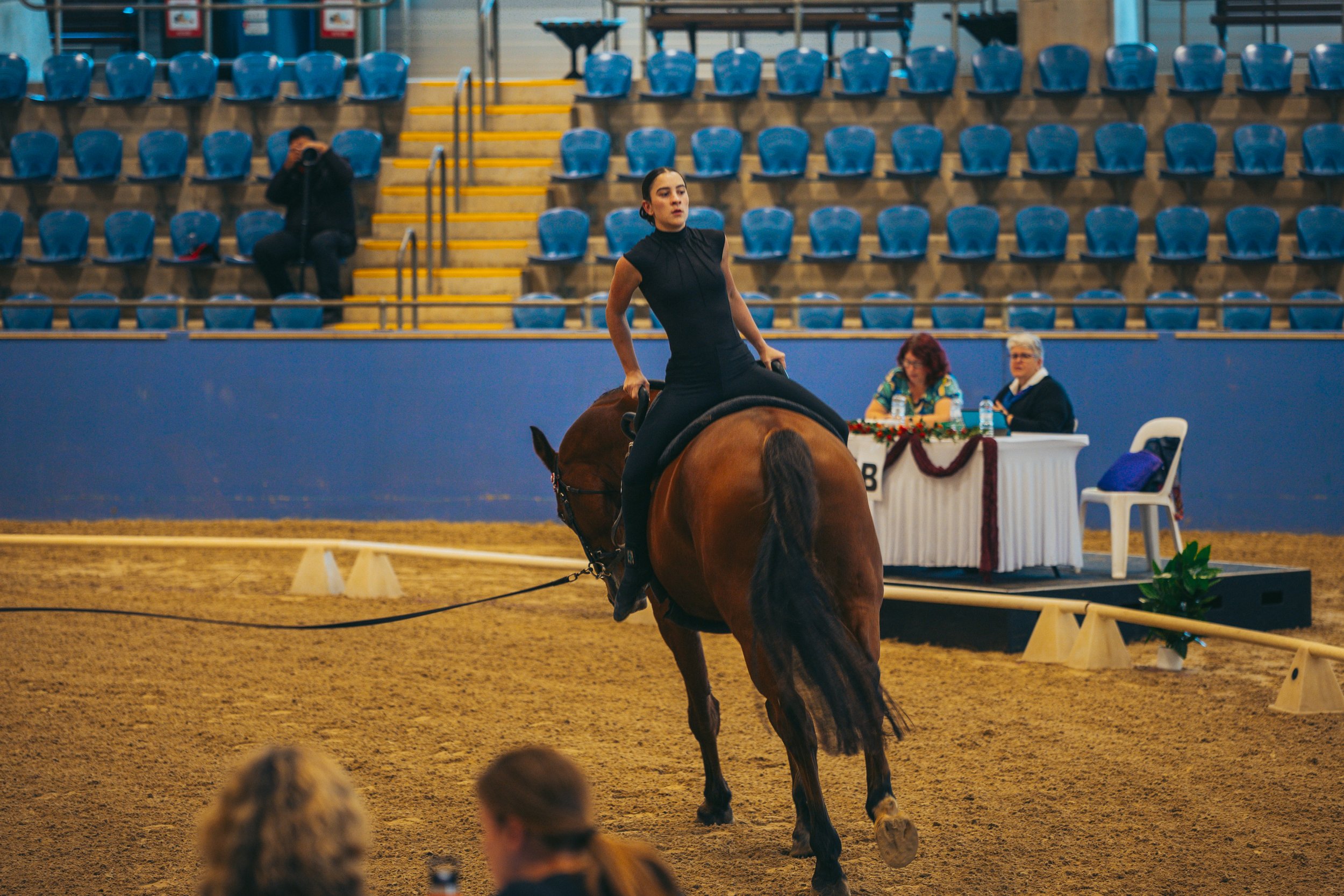 A woman riding a brown horse in an indoor arena, with a judge nearby at a table decorated with holiday garlands and water bottles, and spectators in the background.