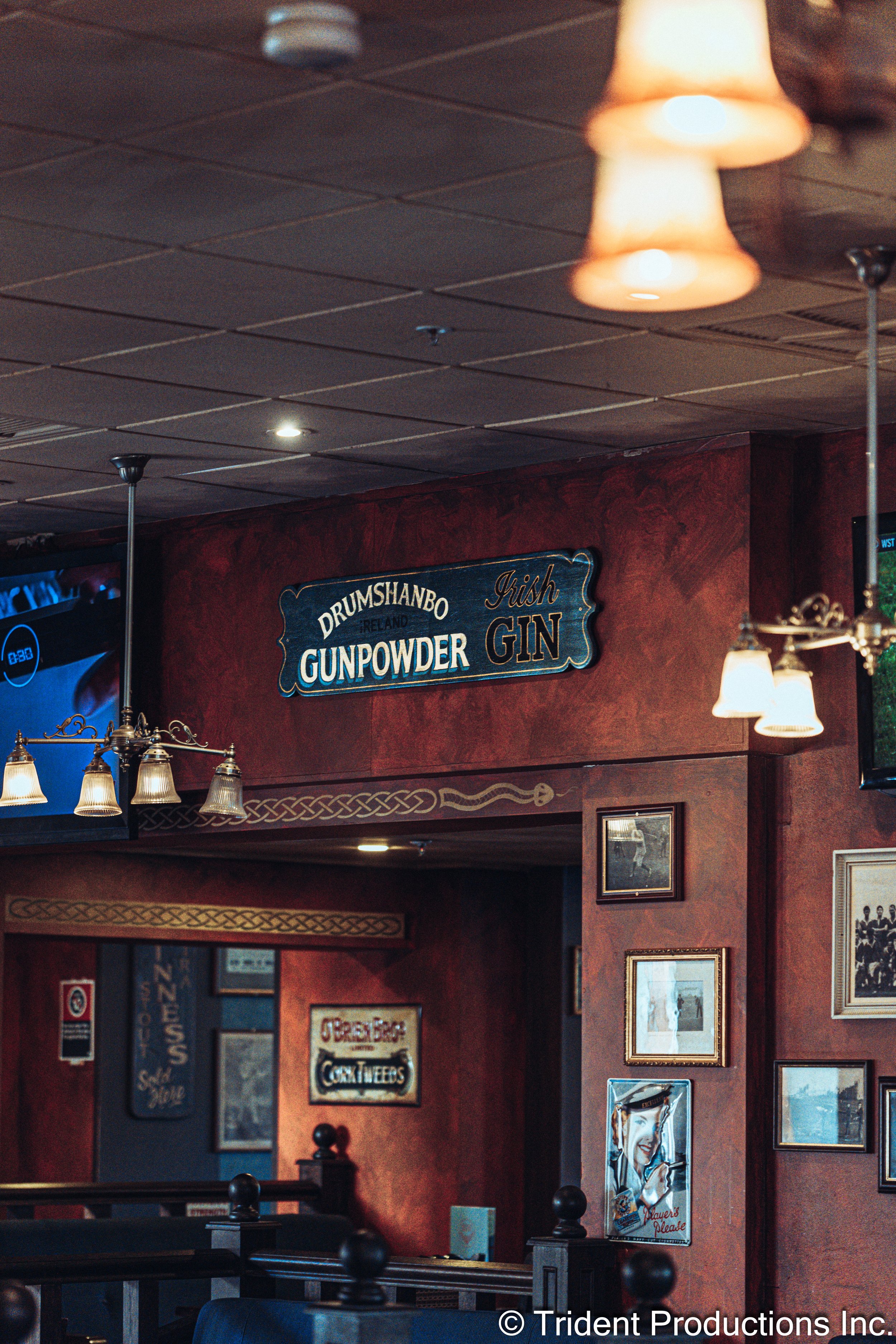 Interior of a bar with a sign that reads 'Drumshanbo Ireland Gunpowder Irish Gin' on a red wall. Decor includes framed pictures, bar lighting fixtures, and a large TV screen.