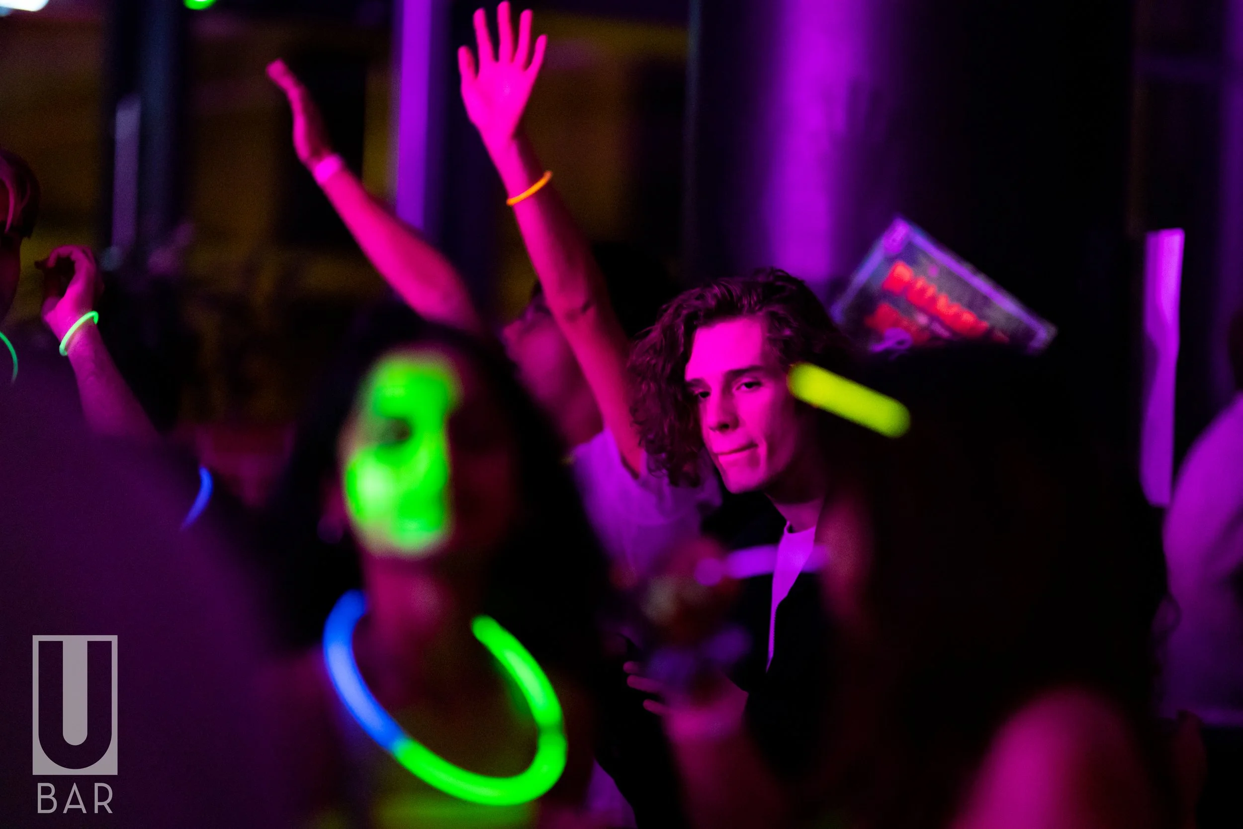 People dancing at a nightclub with colorful neon lights, some wearing glow necklaces and bracelets.