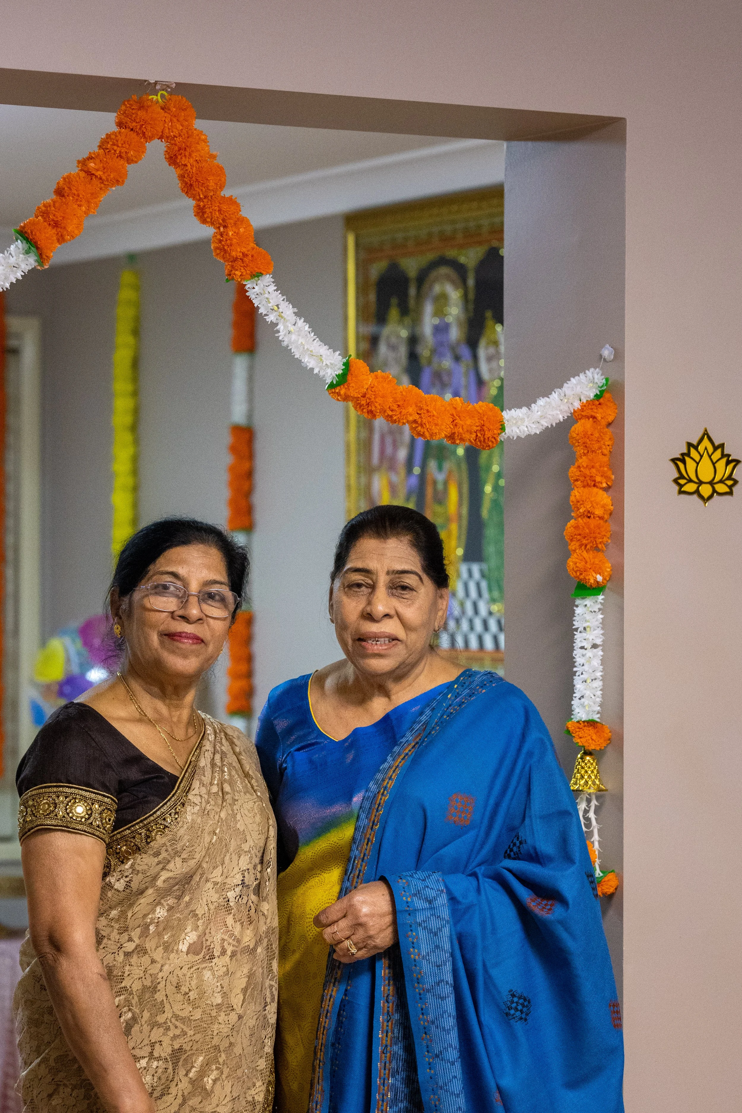 Two women in traditional Indian sarees standing together in a decorated room with orange, white, and green floral garlands.