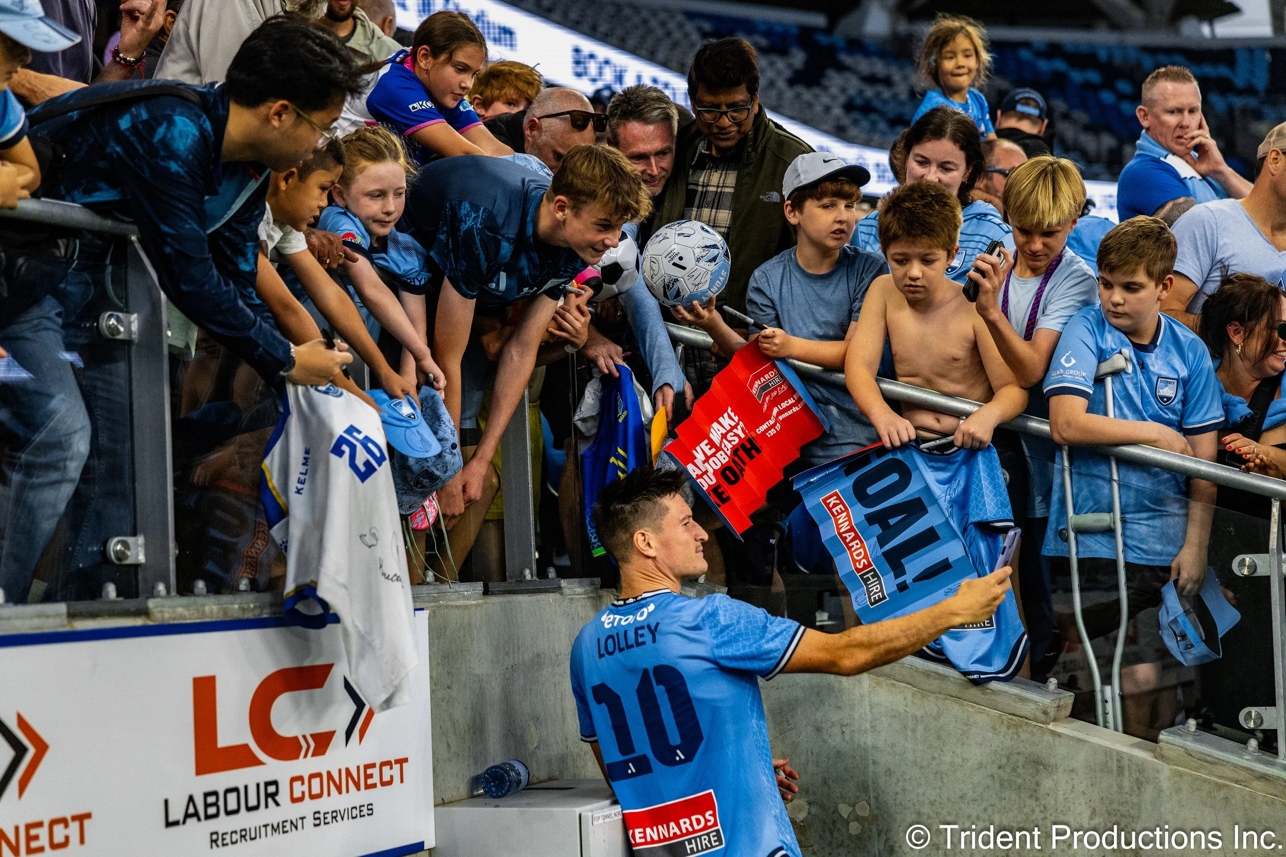Soccer player wearing a light blue jersey signing autographs for enthusiastic young fans at a stadium.