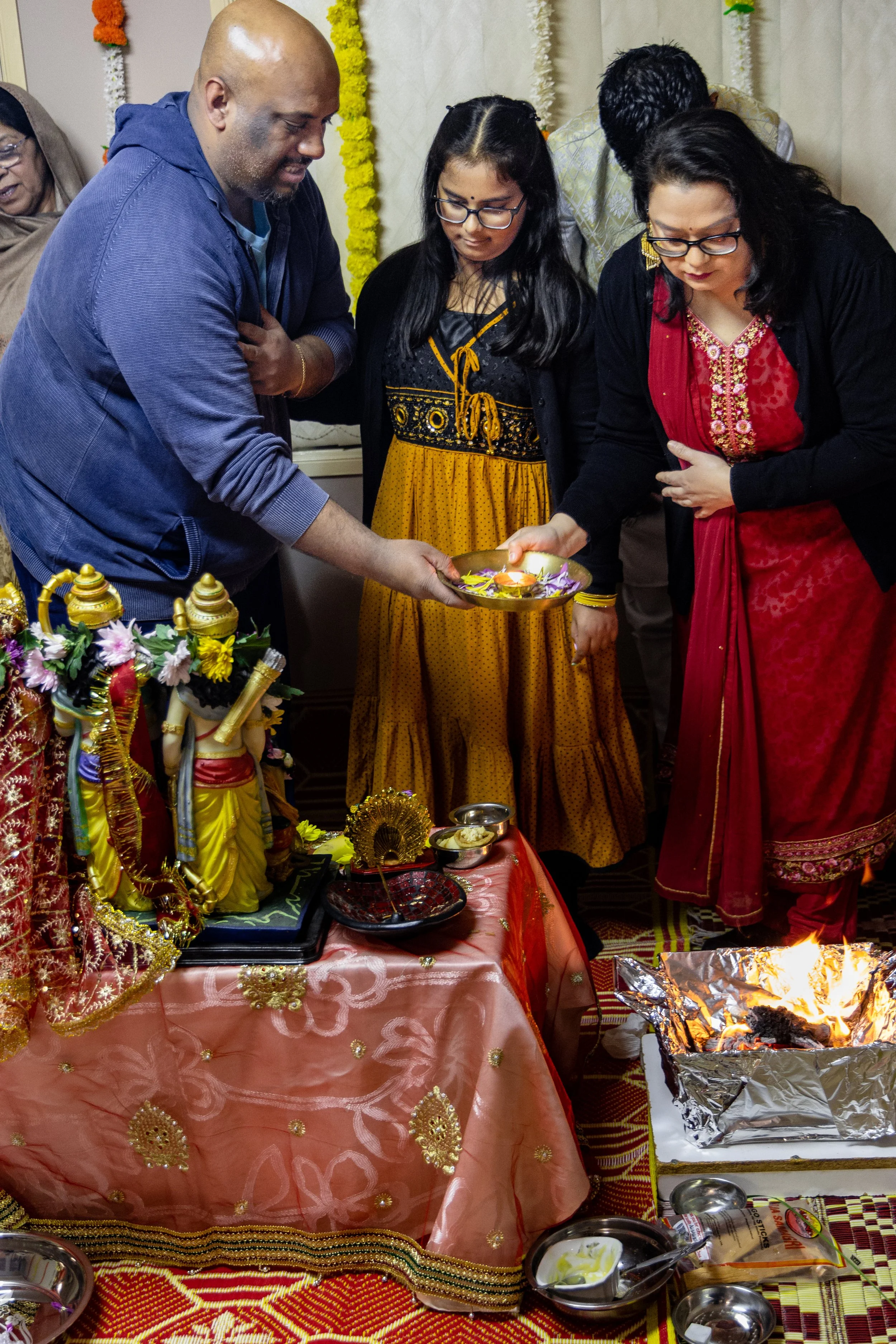 People participating in a Hindu ritual, pouring offerings onto fire, with statues of Hindu deities on a decorated table.