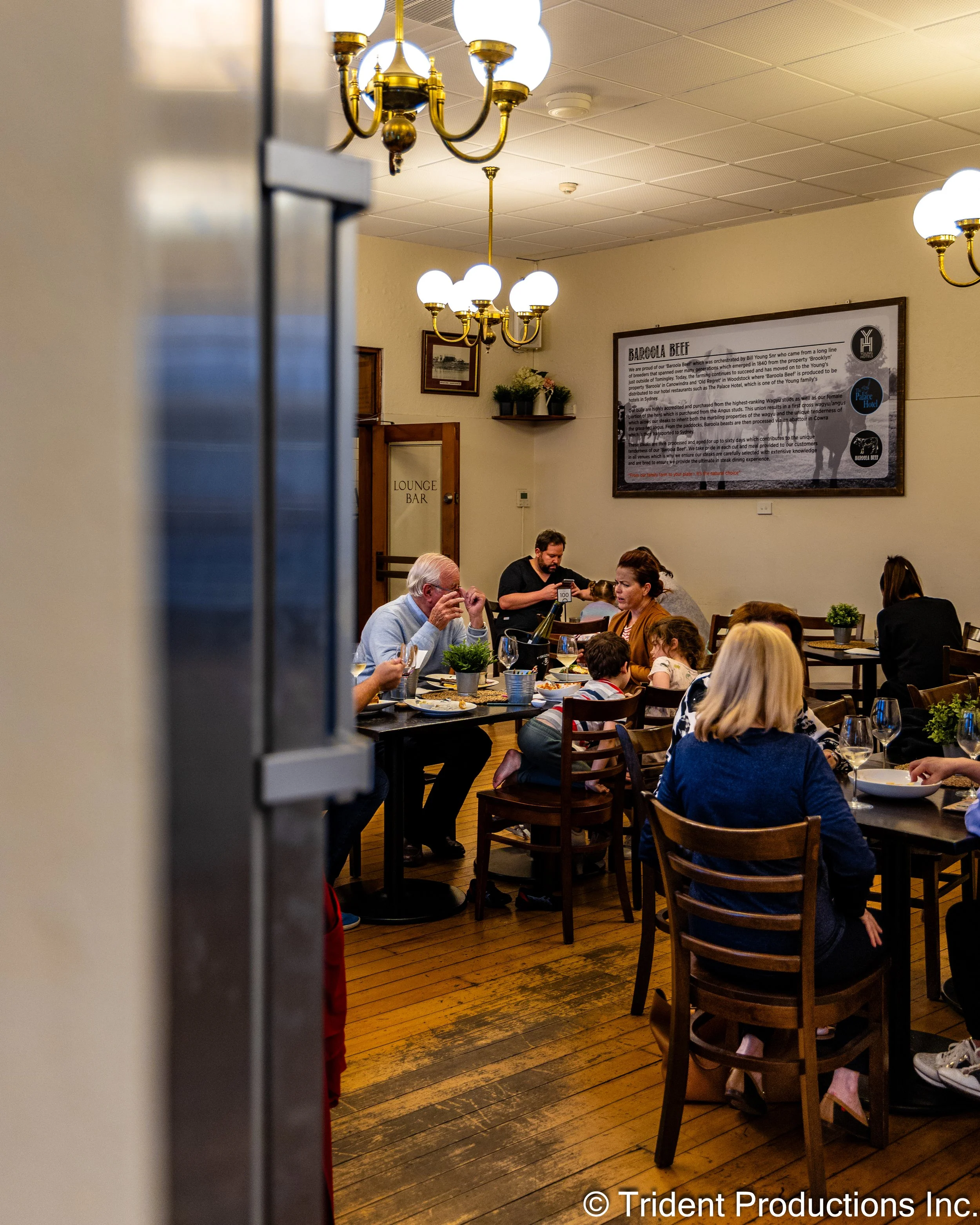 People dining in a cozy restaurant viewed through a partially open door, with chandeliers overhead, a large informational poster on the wall, and warm lighting.