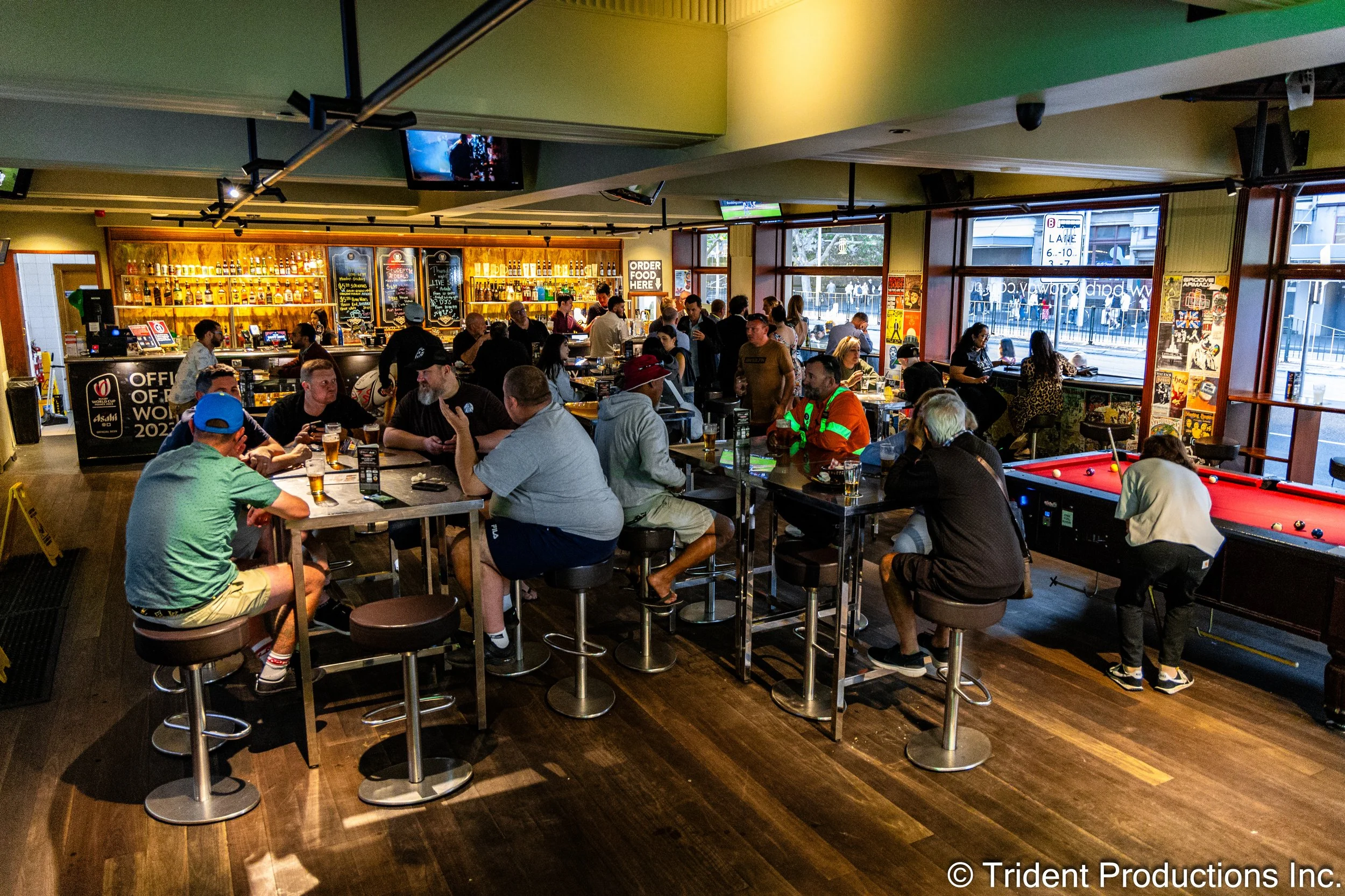 Interior of a lively bar with people sitting at high tables, playing pool, and socializing. Bar shelves stocked with bottles are visible in the background, along with large windows letting in natural light.