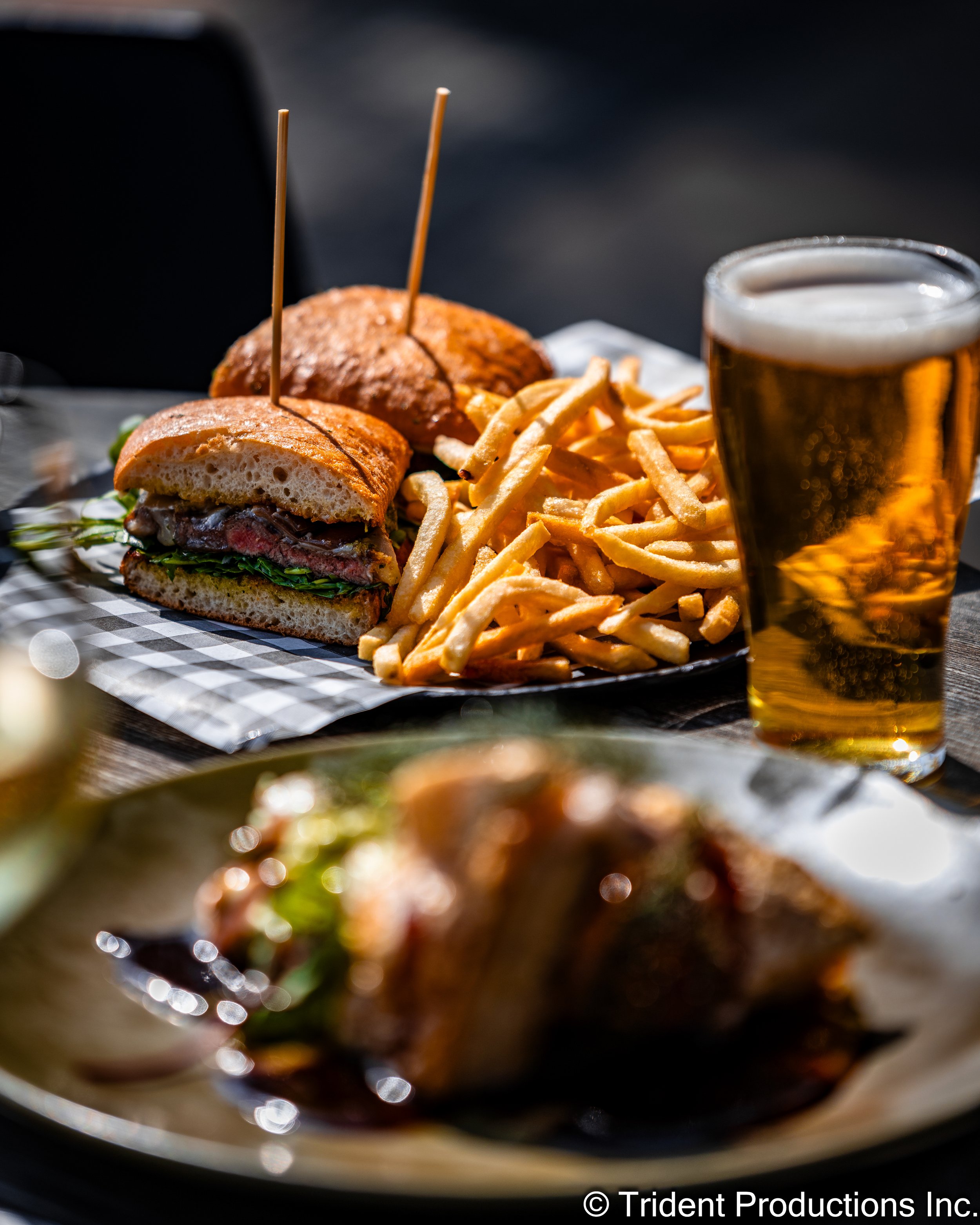 A plate with a cheeseburger and French fries, accompanied by a glass of beer on a dark outdoor table.