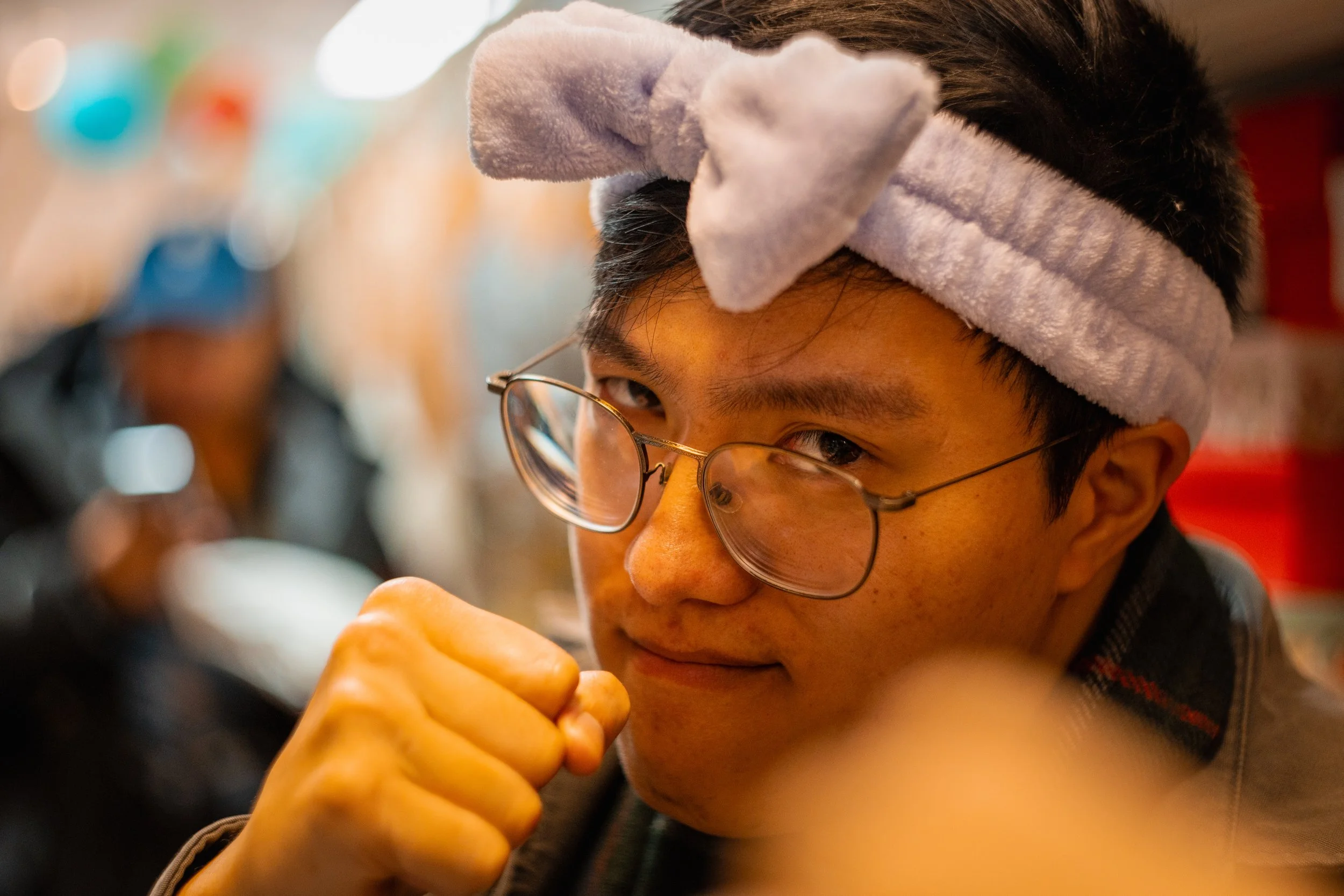 A young man wearing glasses and a fluffy gray headband with a bow, looking at something intently with a slight smile, and resting his chin on his hand in an indoor setting.