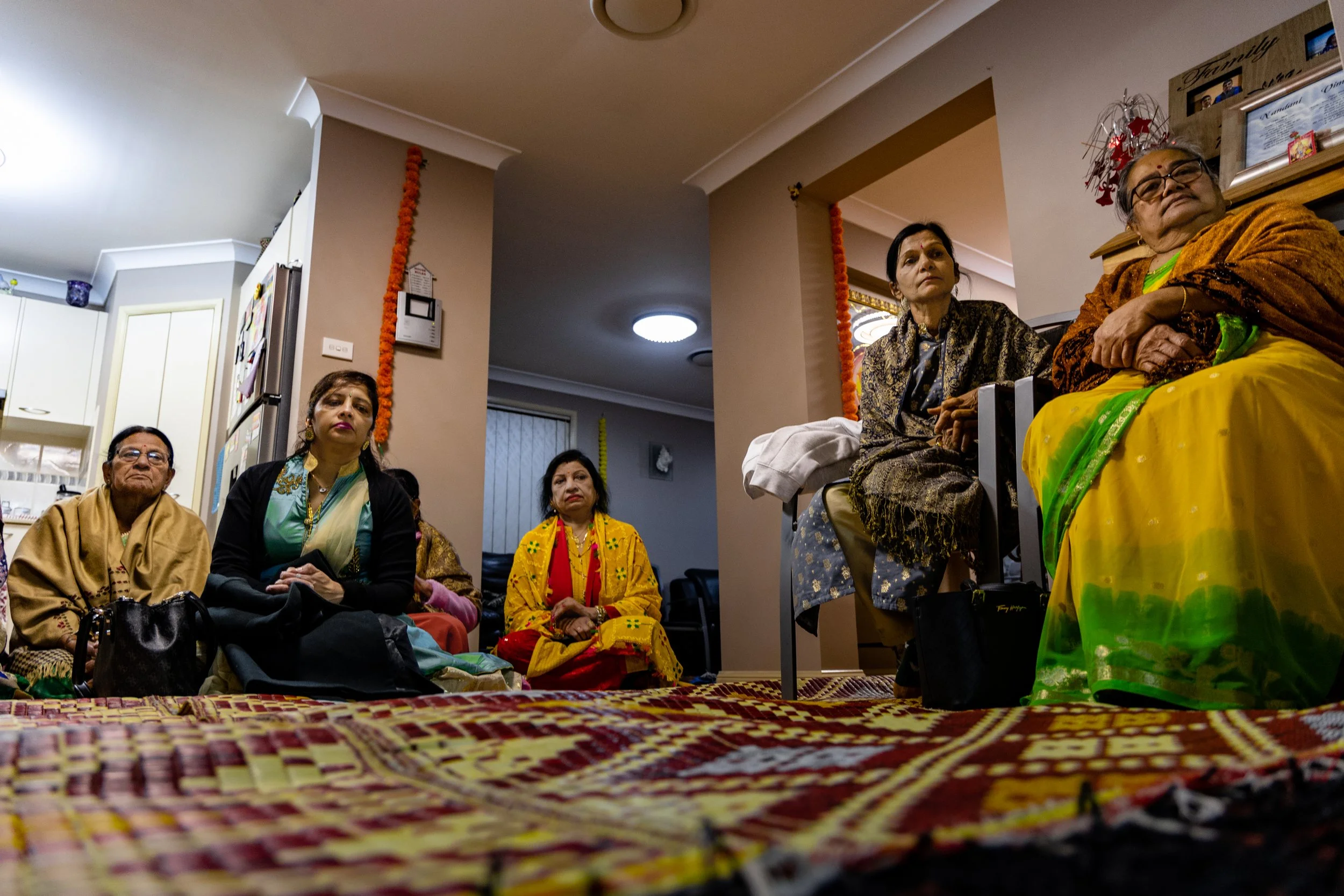 Group of women sitting on the floor and on chairs inside a home, dressed in colorful traditional Indian attire, with some women showing serious or attentive expressions.