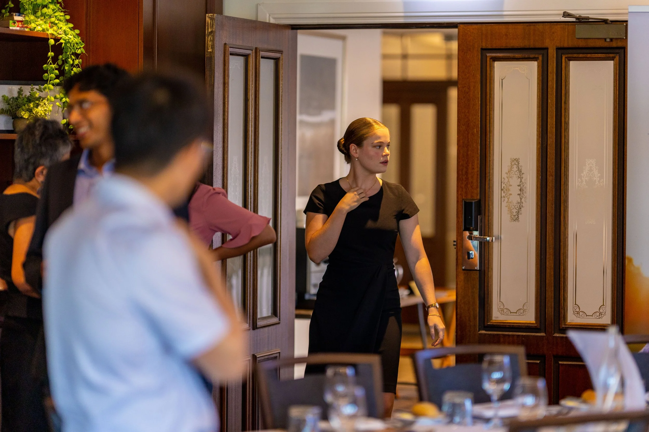 A woman in a black dress standing in a doorway, looking to the side, at a formal event with tables and glasses in the foreground, and other people in the background.