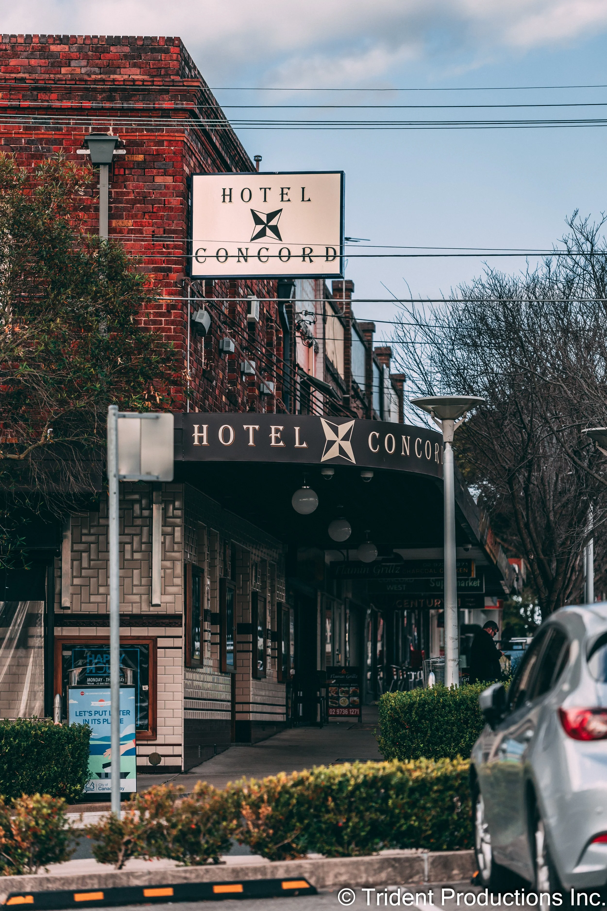 Street view of Hotel Concord with a sign displaying the hotel name. The hotel building has a brick facade and a canopy over the sidewalk. There are trees, street lamps, parked cars, and a person walking on the sidewalk.