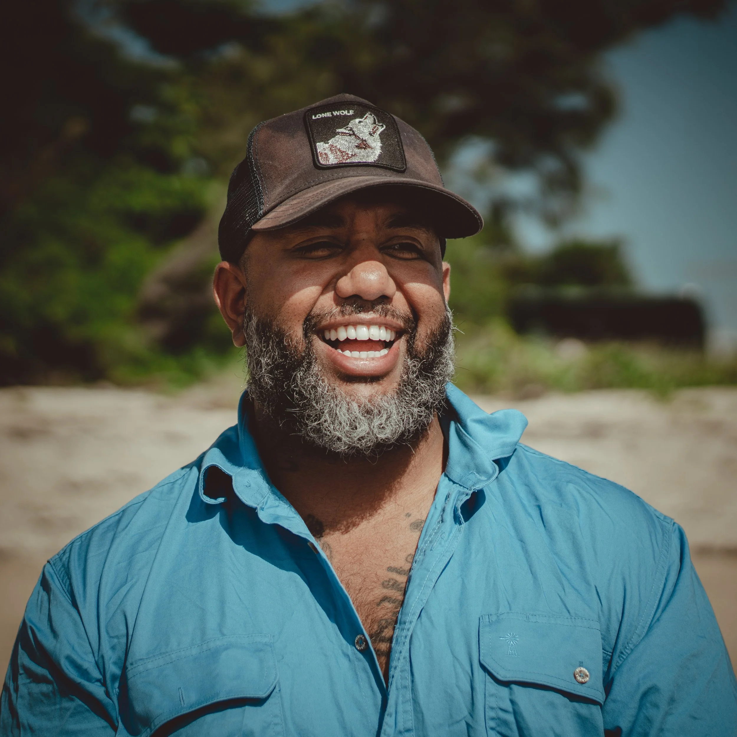 A man with a gray beard smiling on a beach, wearing a blue shirt and a brown cap with a wolf patch.